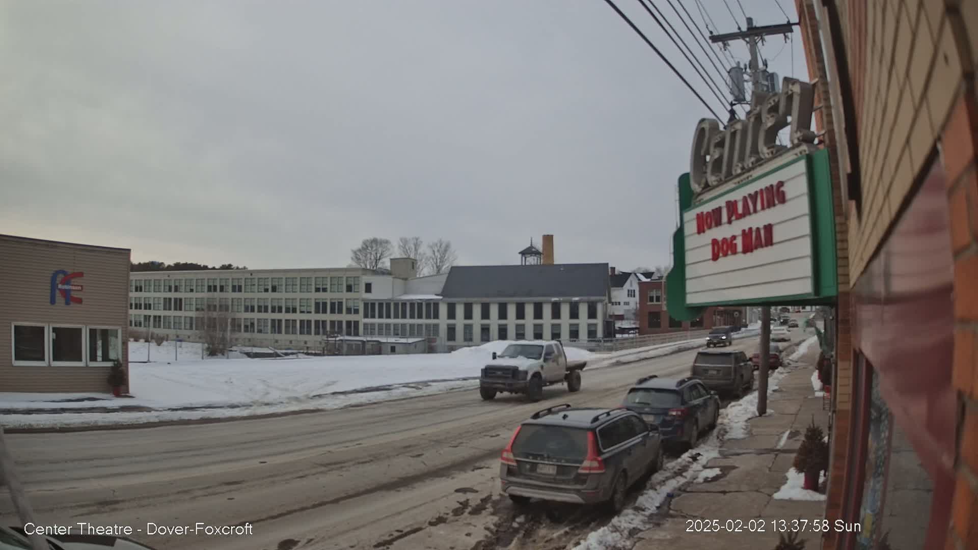 A snow-covered street scene shows several cars parked along a curb near a movie theater marquee, with larger buildings and a snowy, overcast sky in the background.