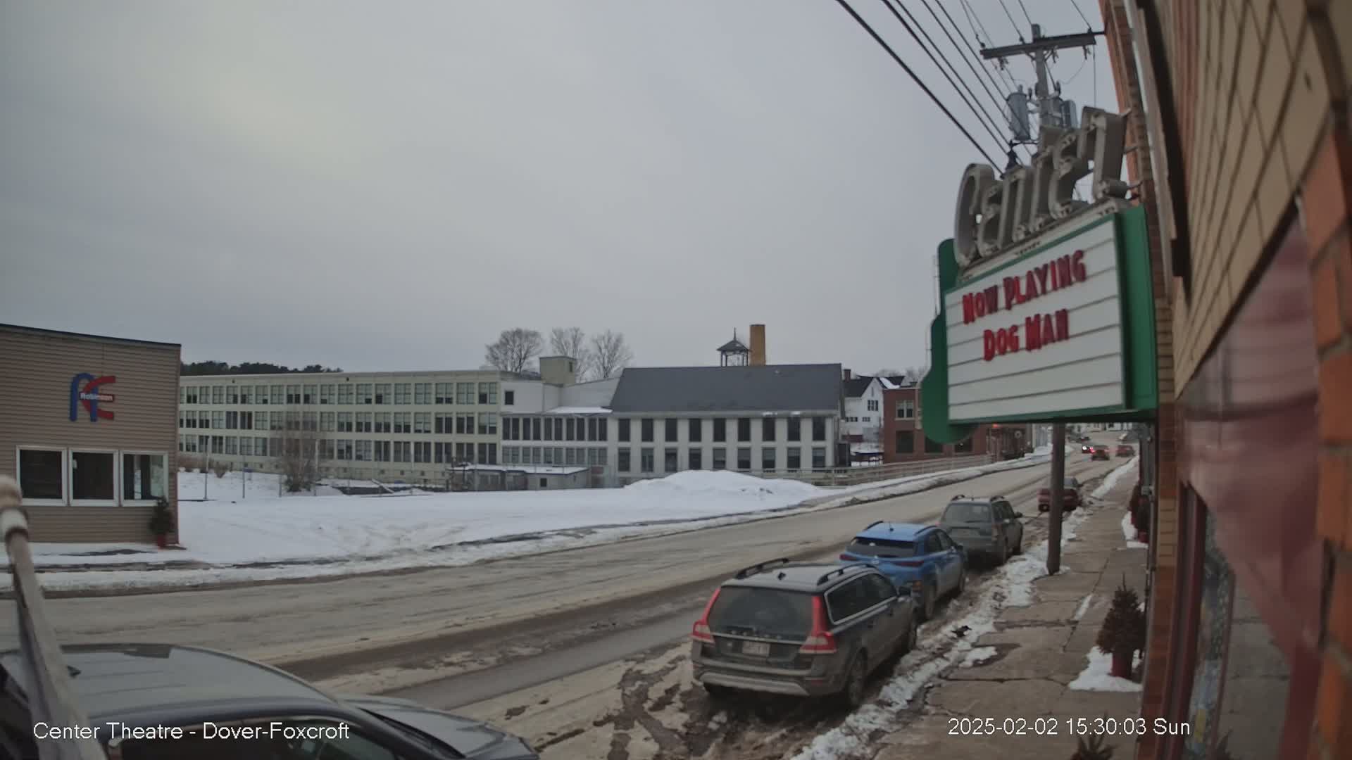 A snow-covered street scene with cars parked along the side, several buildings in the background, and overcast weather.