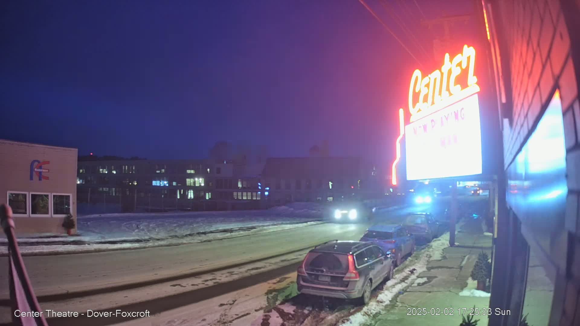 A snow-covered street at night, with cars parked along the side and a brightly lit sign for a theater in the background.