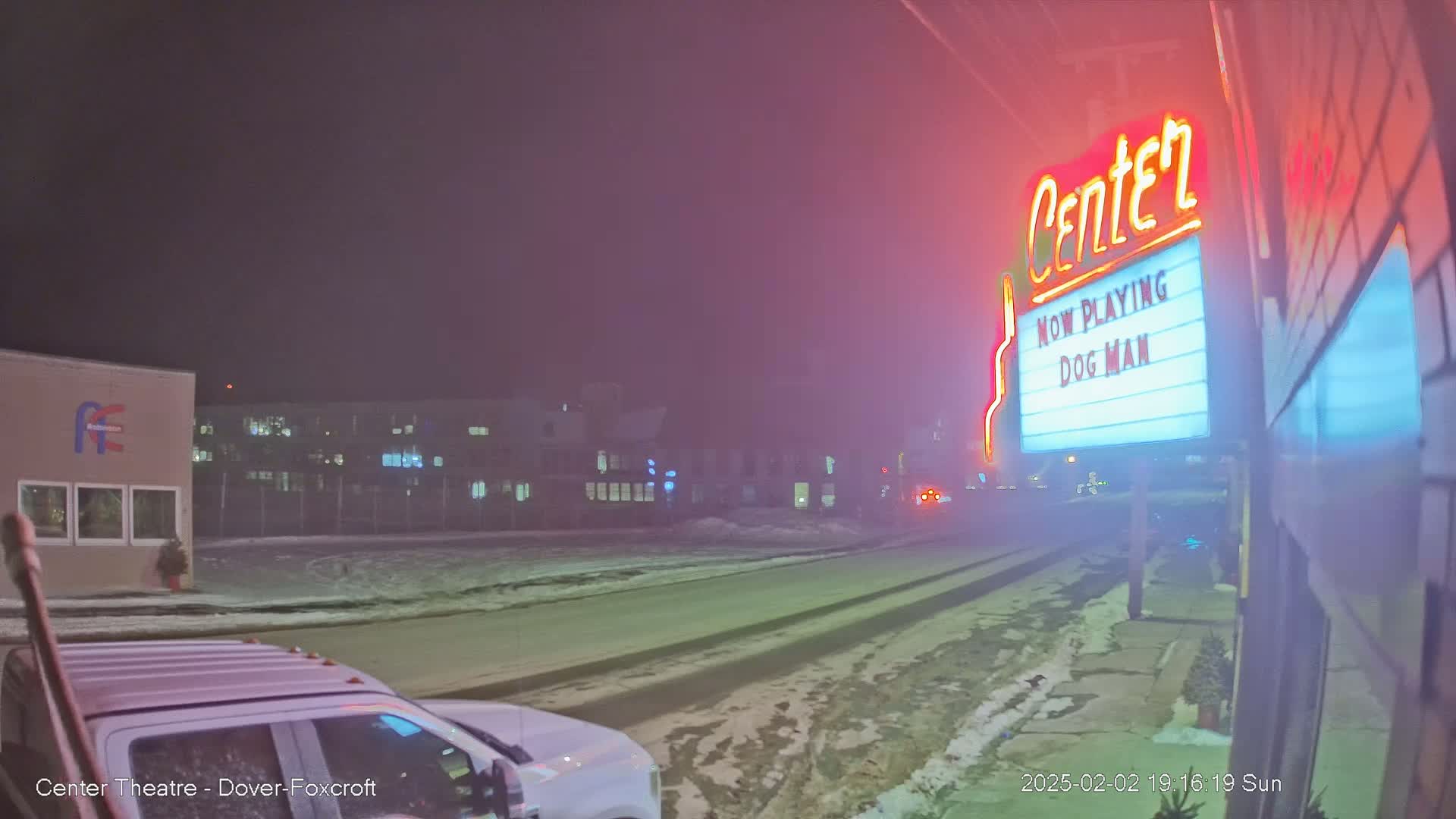 A snowy, foggy night scene shows a movie theater marquee advertising "Dog Man," a street with a vehicle partially visible, and buildings in the background.