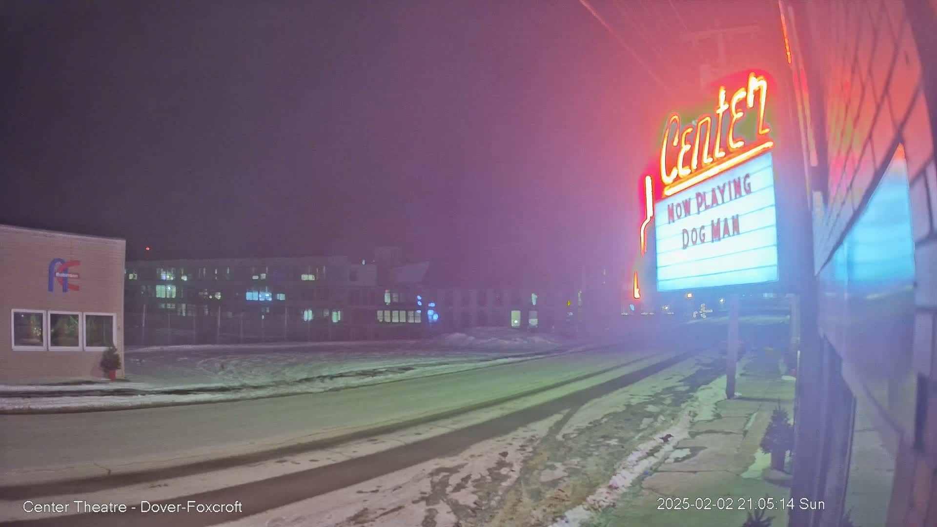 A foggy, snowy night scene shows a movie theater marquee advertising "Dog Man," with a road and buildings visible in the background.