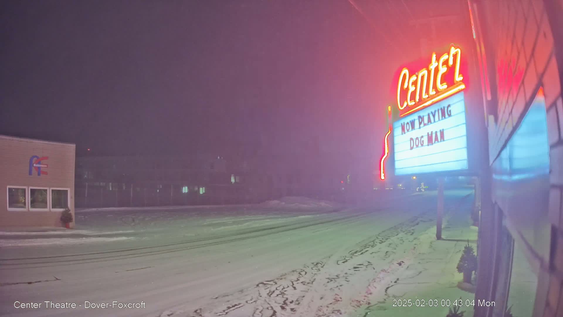 A snow-covered street at night is seen with a movie theater marquee displaying "Dog Man" in the foreground, under a hazy, pinkish sky.