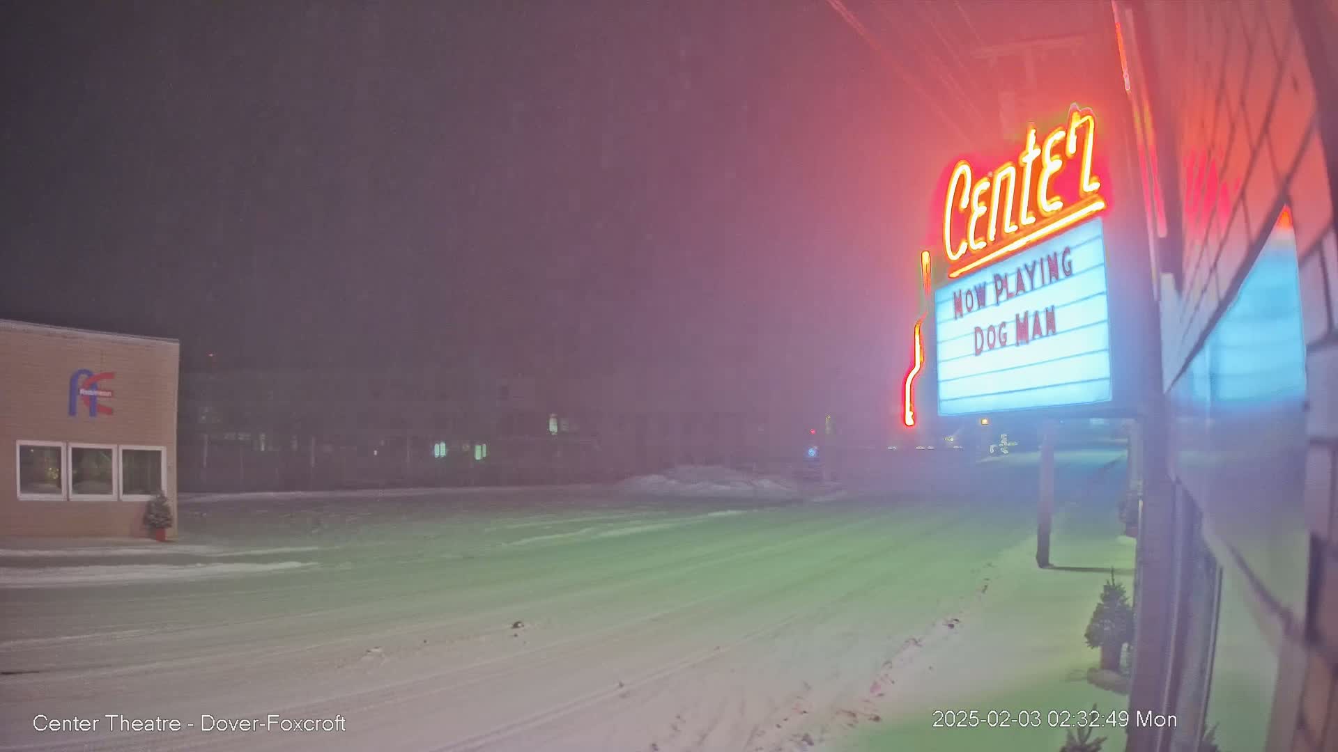 A snow-covered street at night, with a movie theater marquee advertising "Dog Man" illuminated by neon lights in the foreground.
