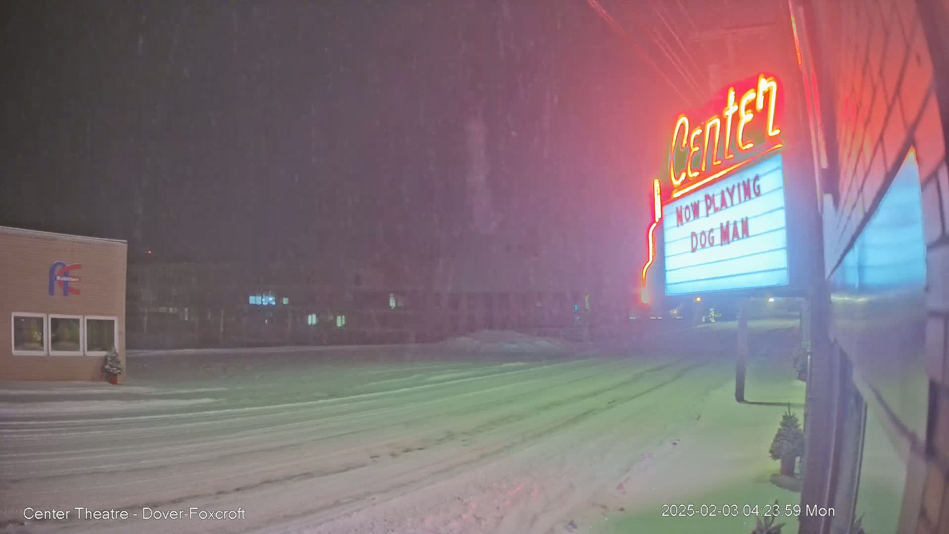 A snow-covered street at night, with a movie theater marquee displaying "Dog Man" visible in the falling snow.