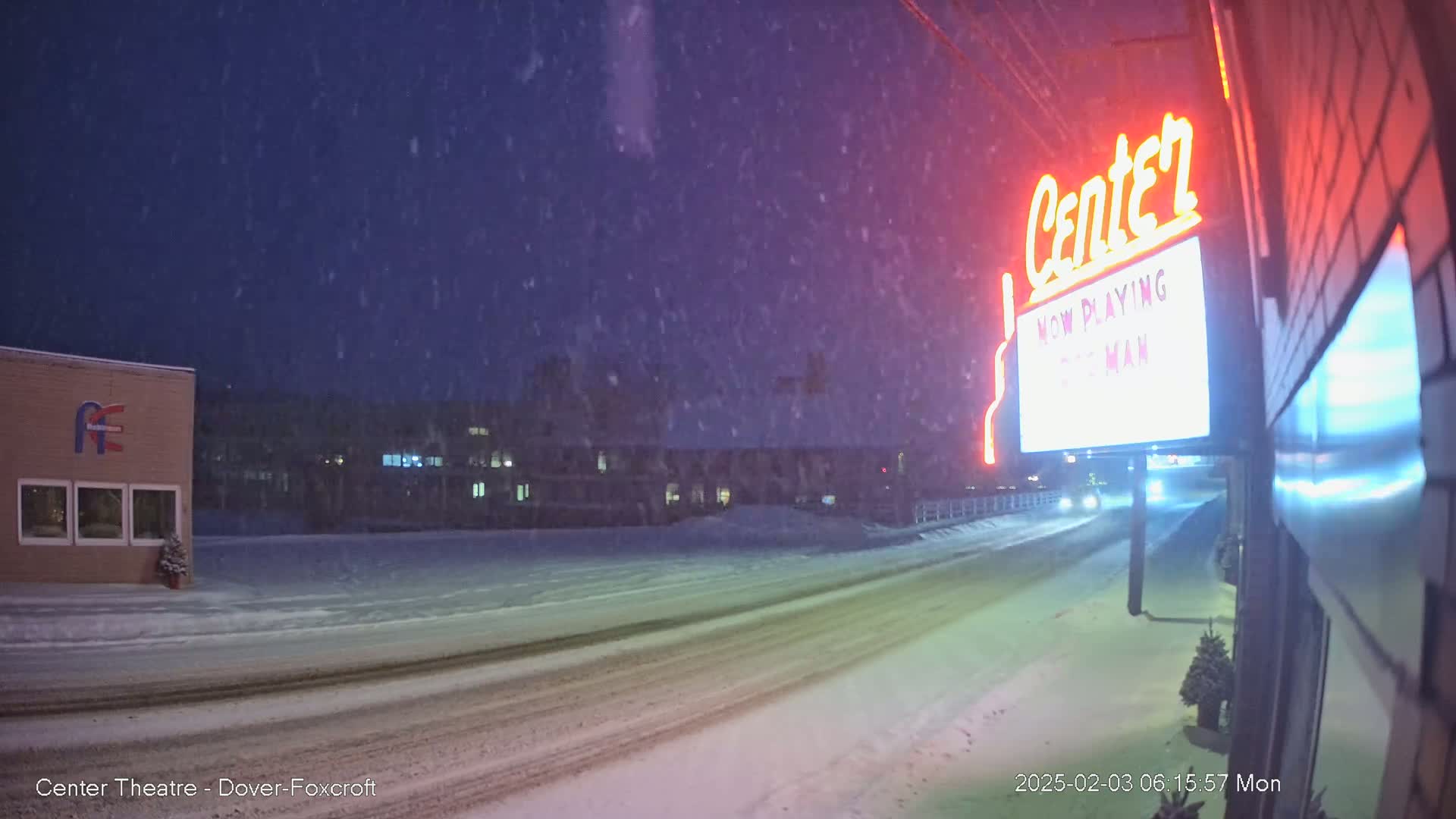 A snow-covered street at night is seen with a movie theater marquee displaying "NOW PLAYING" and a partially visible movie title, with some buildings in the background under a falling snow.