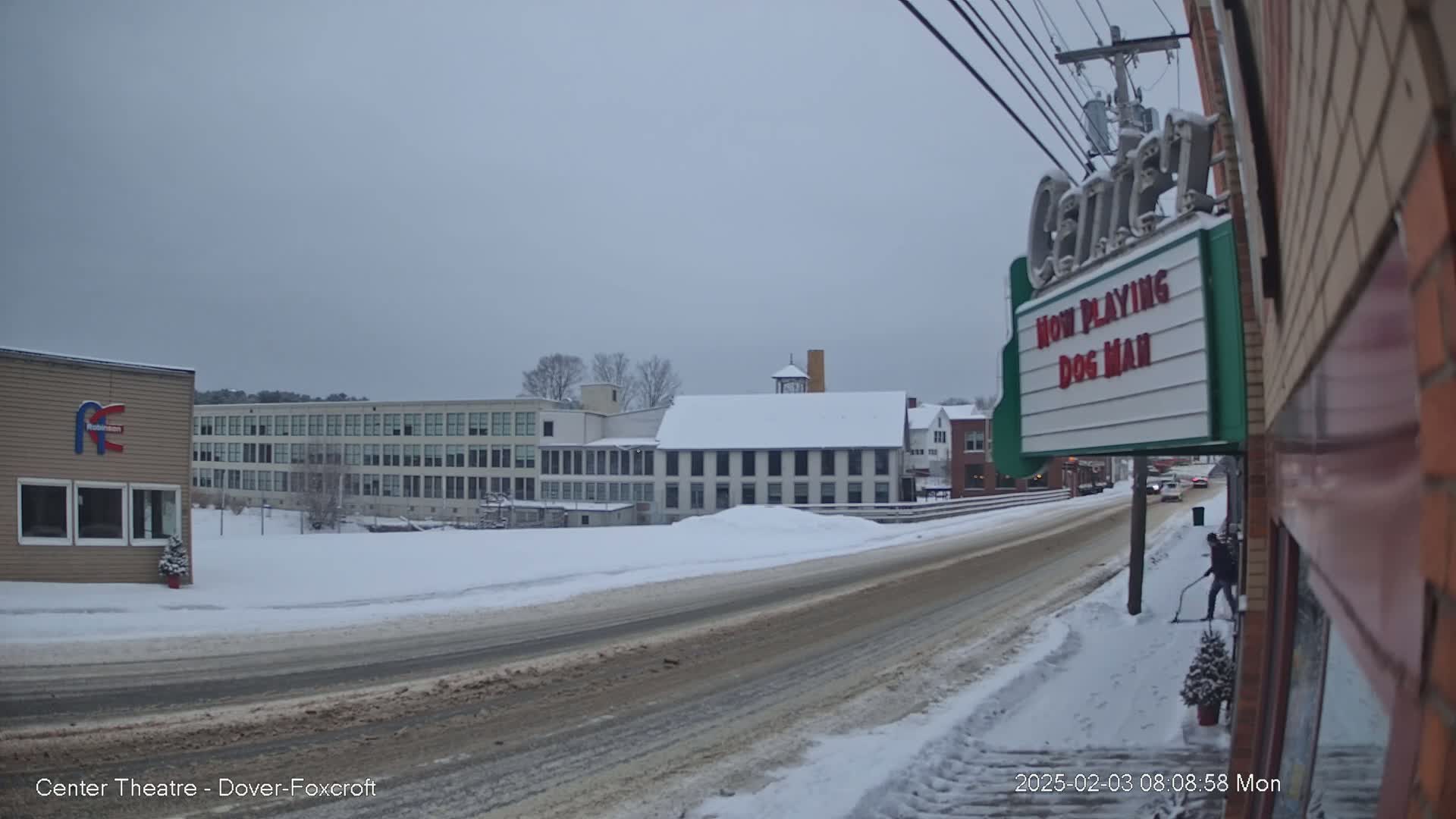 A snow-covered street scene shows several buildings, including a large factory and a movie theater marquee advertising "Dog Man," under an overcast sky.