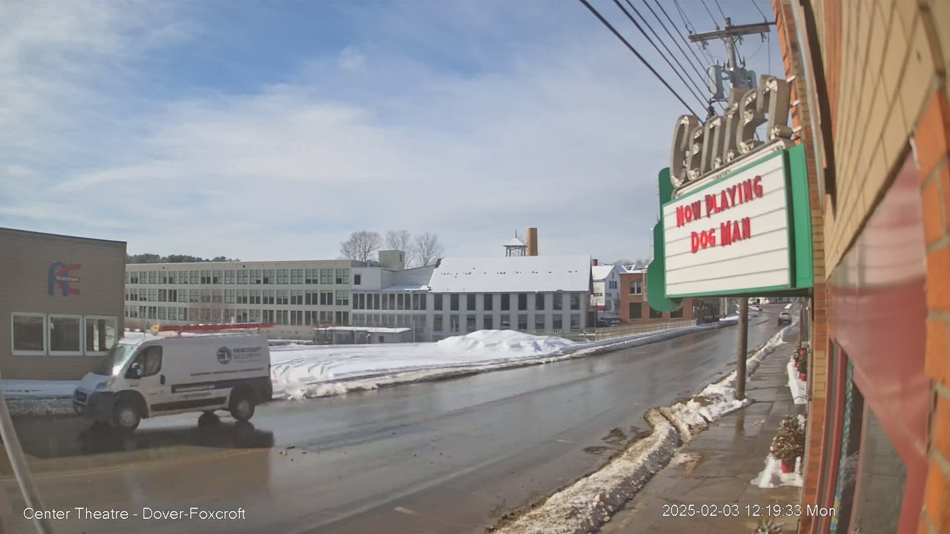 A mostly sunny day reveals a street scene with snow-covered patches along the sides, a wet road, several buildings including a multi-story factory, and a white van driving.