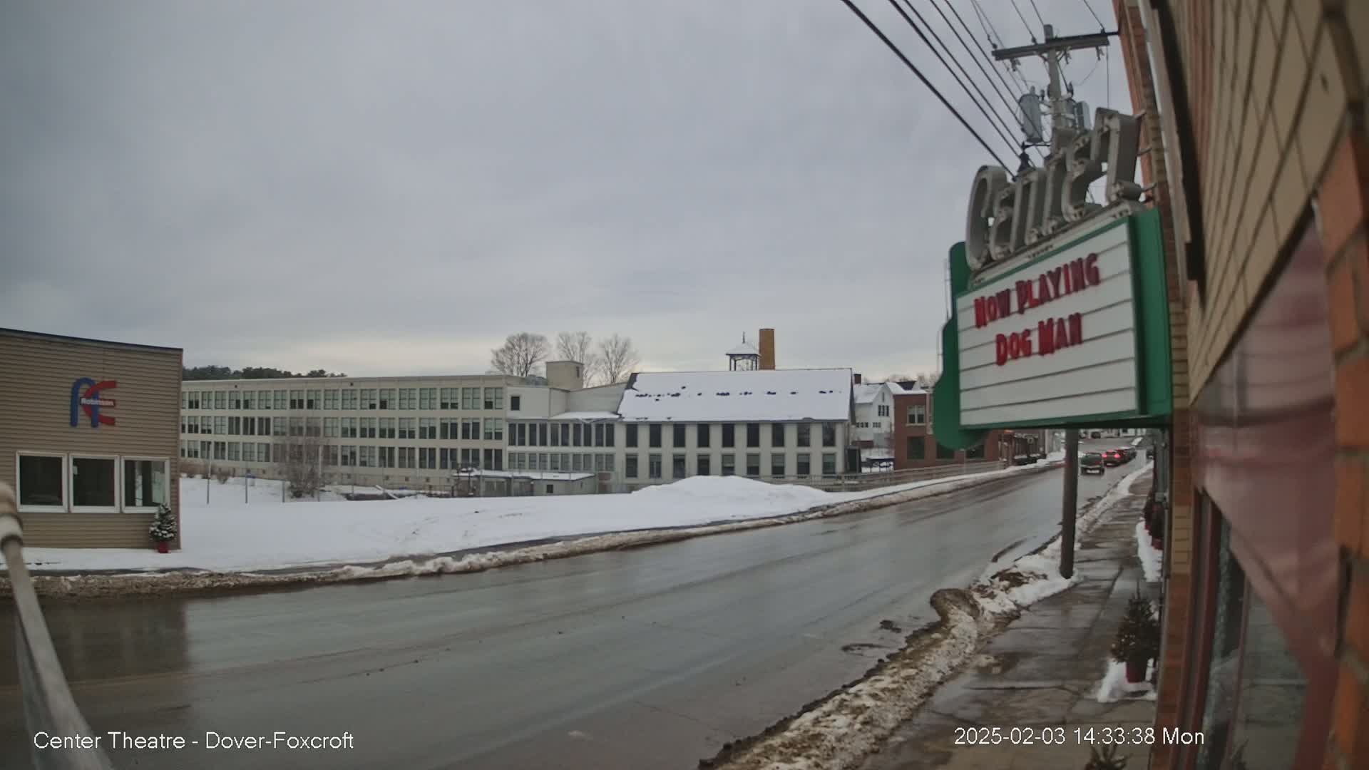 A wet road on a cloudy day passes several buildings, including a large multi-story building and a movie theater marquee that shows “Dog Man” is playing.