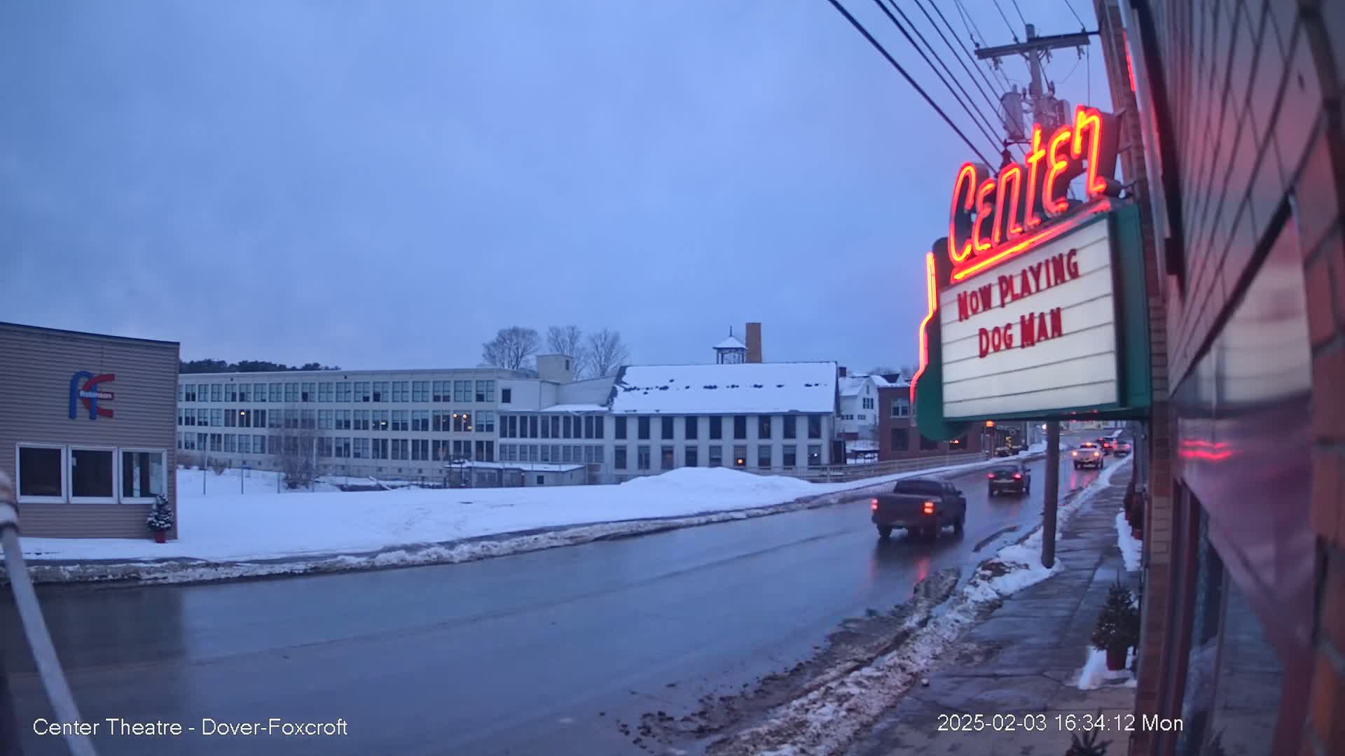 A snow-covered street scene at dusk shows a movie theater marquee, several buildings, and a few cars driving on a wet road.