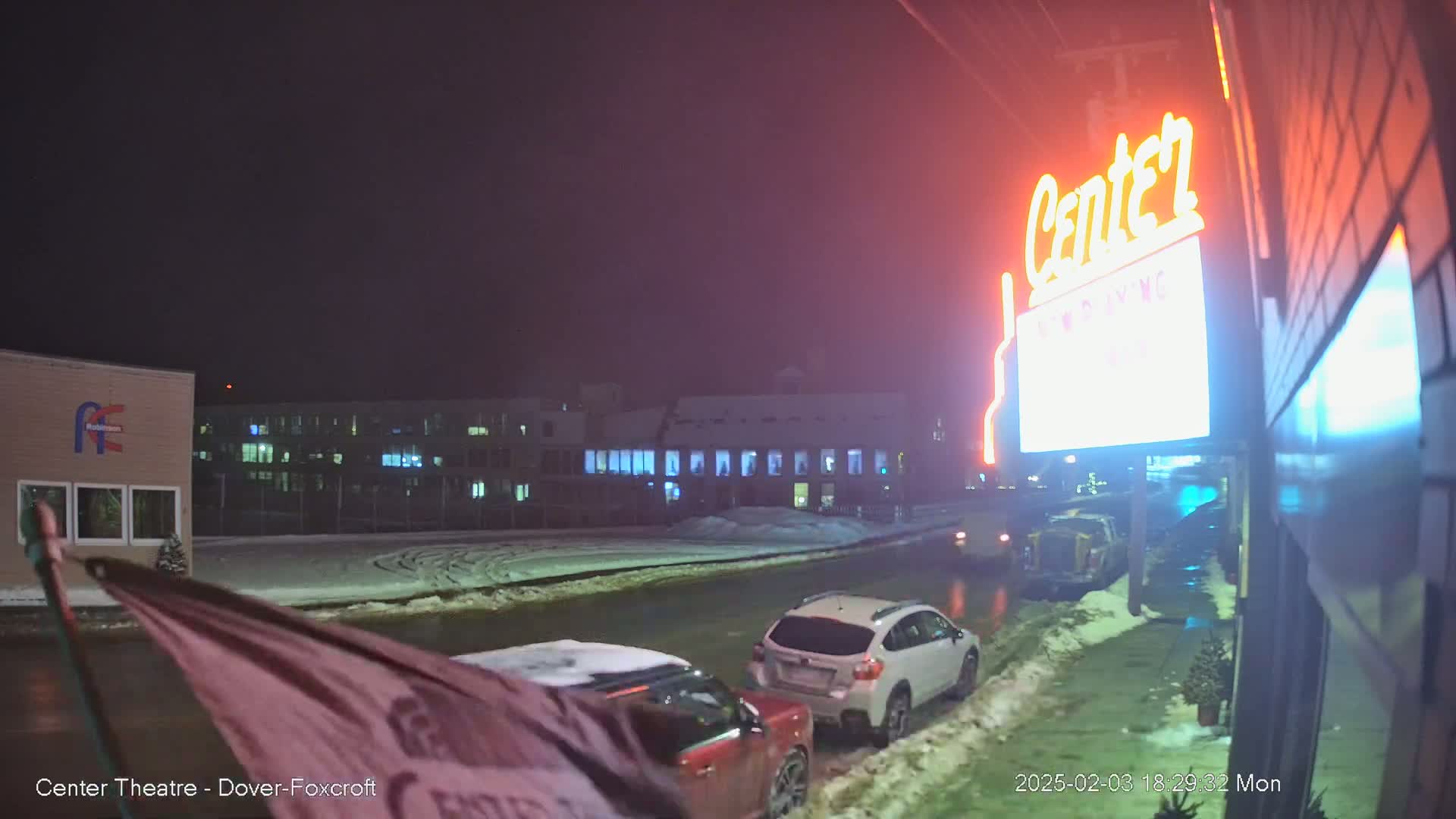 A nighttime scene shows a snow-covered parking lot with cars parked, a building with a lit-up sign that says "Center", and other buildings in the background under a dark, overcast sky.