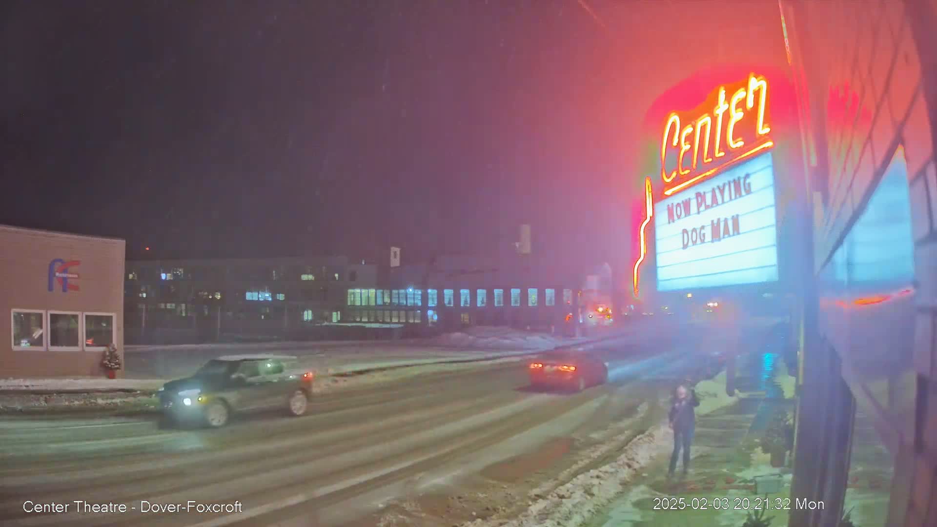 A snowy night scene shows a street with cars driving past a movie theater marquee advertising "Dog Man."