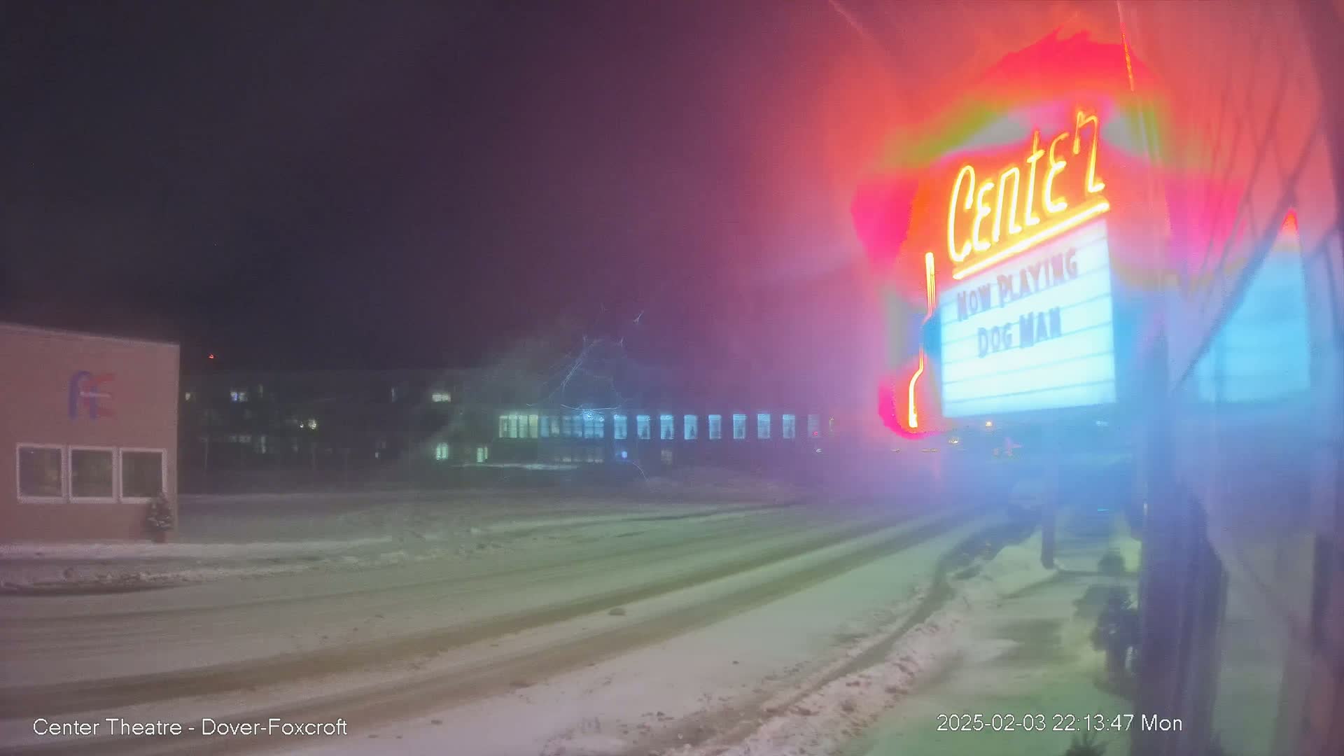 A snow-covered street at night is seen with a brightly lit movie theater marquee in the foreground showing "Dog Man" is now playing.