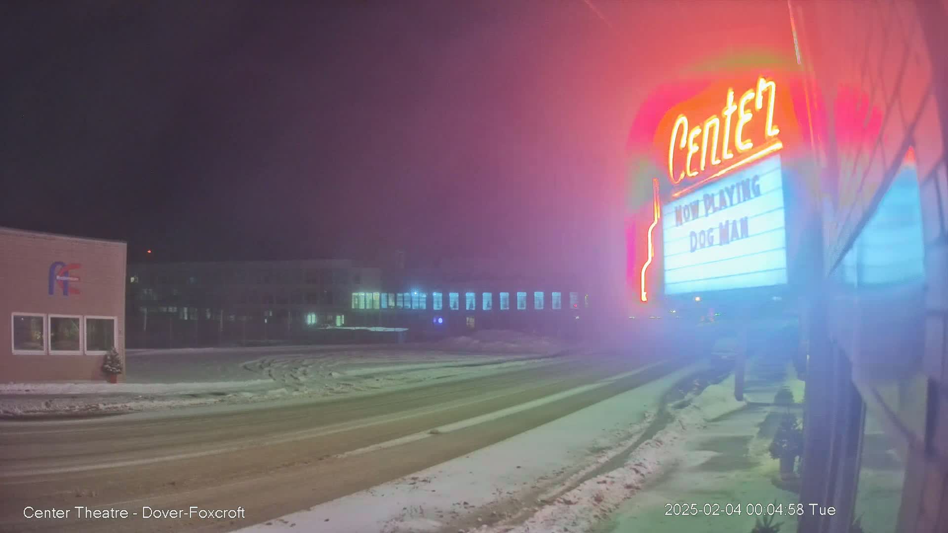 A snow-covered street at night is seen in front of a movie theater marquee displaying "Dog Man," with a hazy, foggy atmosphere.