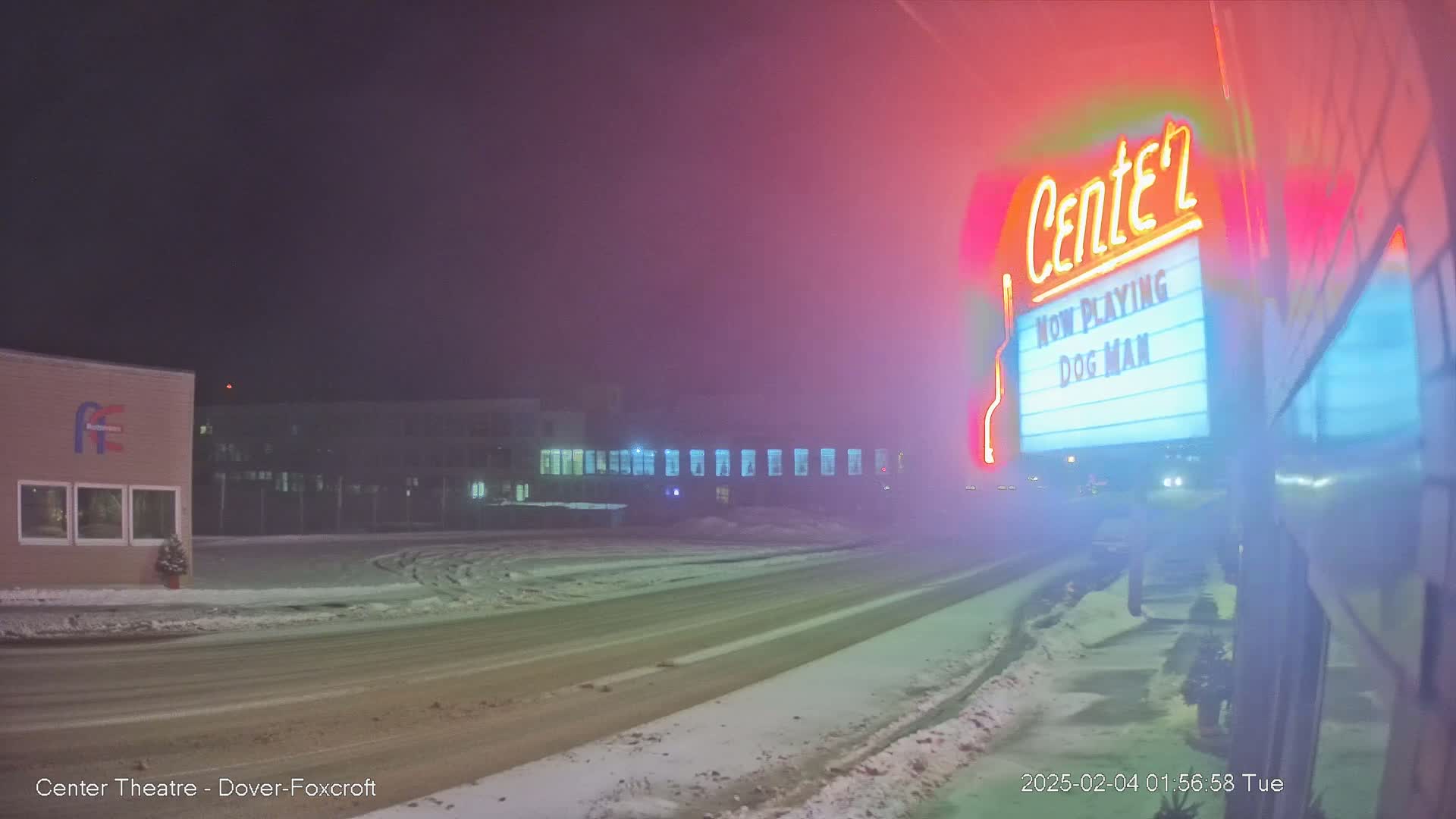 A snow-covered street at night, dimly lit, with a movie theater marquee advertising "Dog Man" visible through a foggy atmosphere.