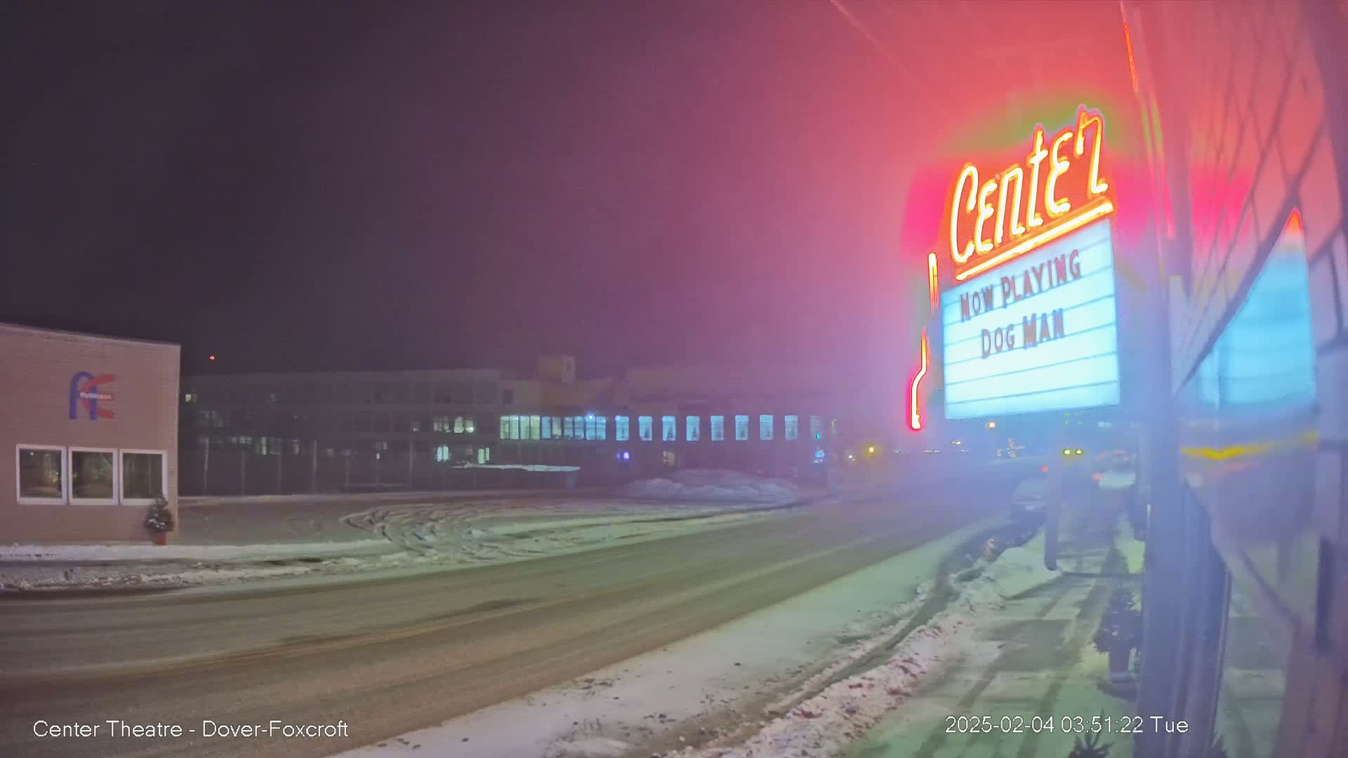 A snow-covered street at night leads to a movie theater marquee advertising "Dog Man" under foggy conditions.
