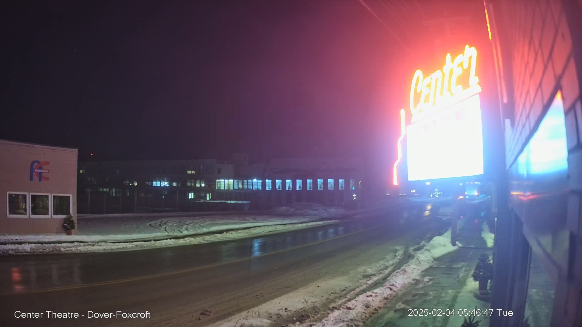 A wet road at night, bordered by snow, leads to a building with illuminated windows and a neon sign.
