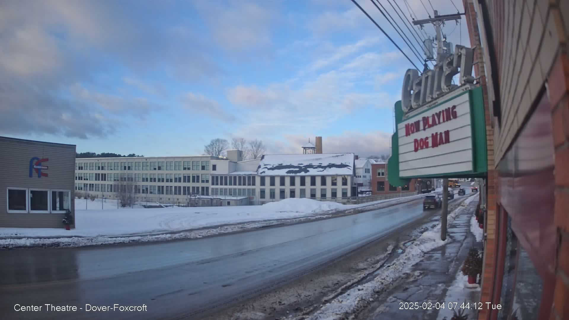 A mostly clear, cold day shows a snow-covered street scene with buildings, including a movie theater marquee advertising "Dog Man," and several cars driving on a wet road.