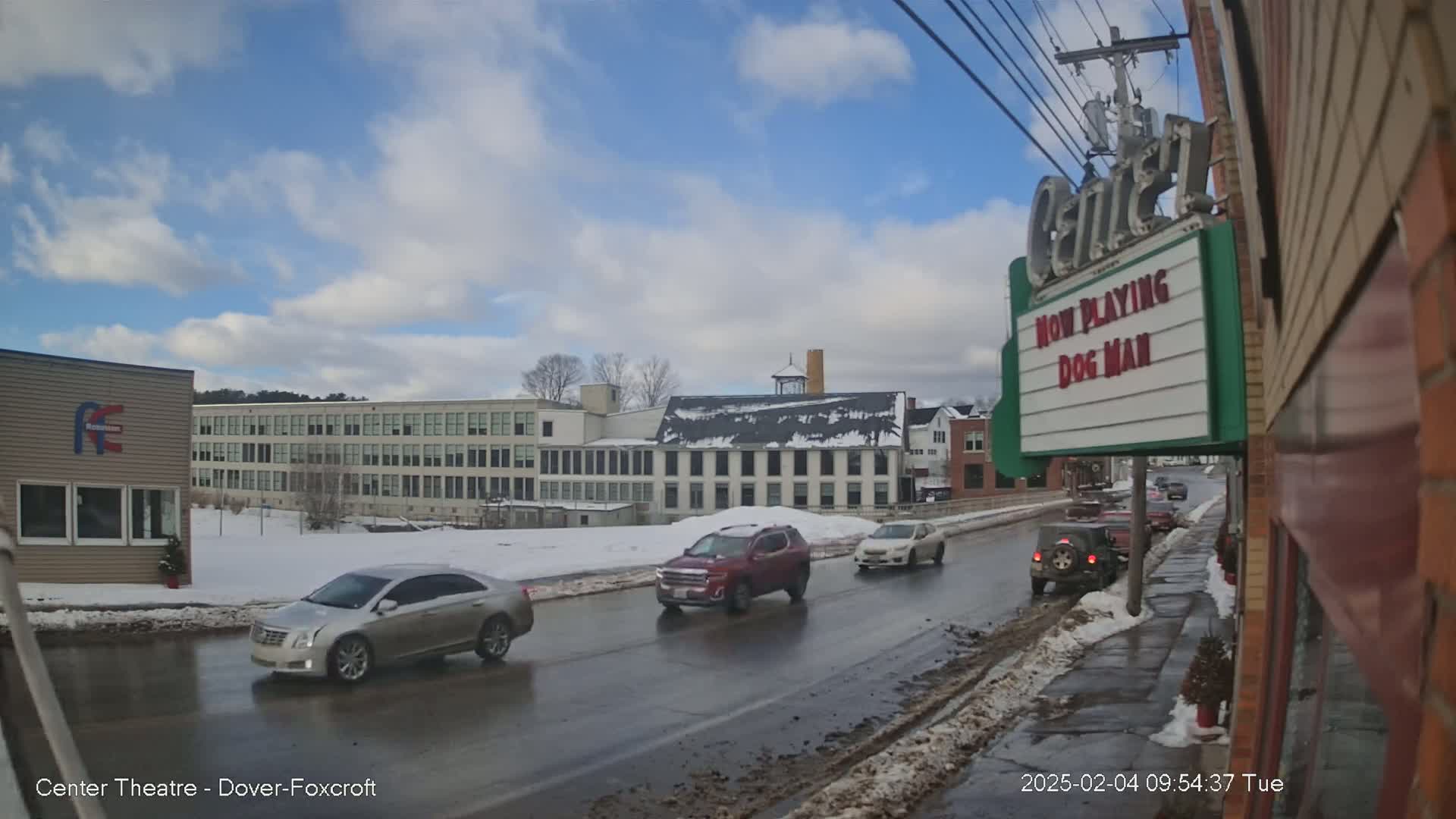 A partly cloudy day reveals a street scene with several cars driving past a multi-story building and a movie theater marquee displaying "Dog Man."