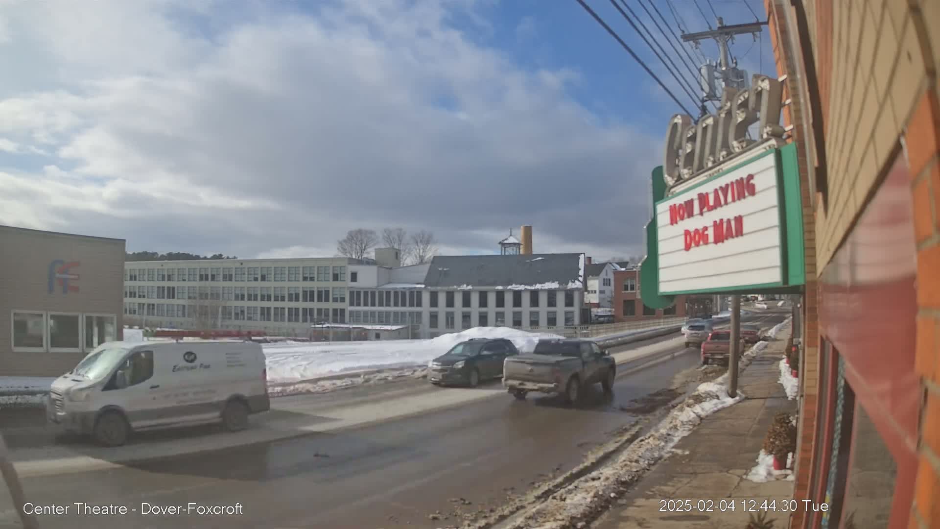 A partly cloudy day reveals a street scene with several vehicles, including a van, an SUV, and a pickup truck, driving past buildings, one of which is a movie theater showing "Dog Man," with snow visible on the ground and sides of the road.