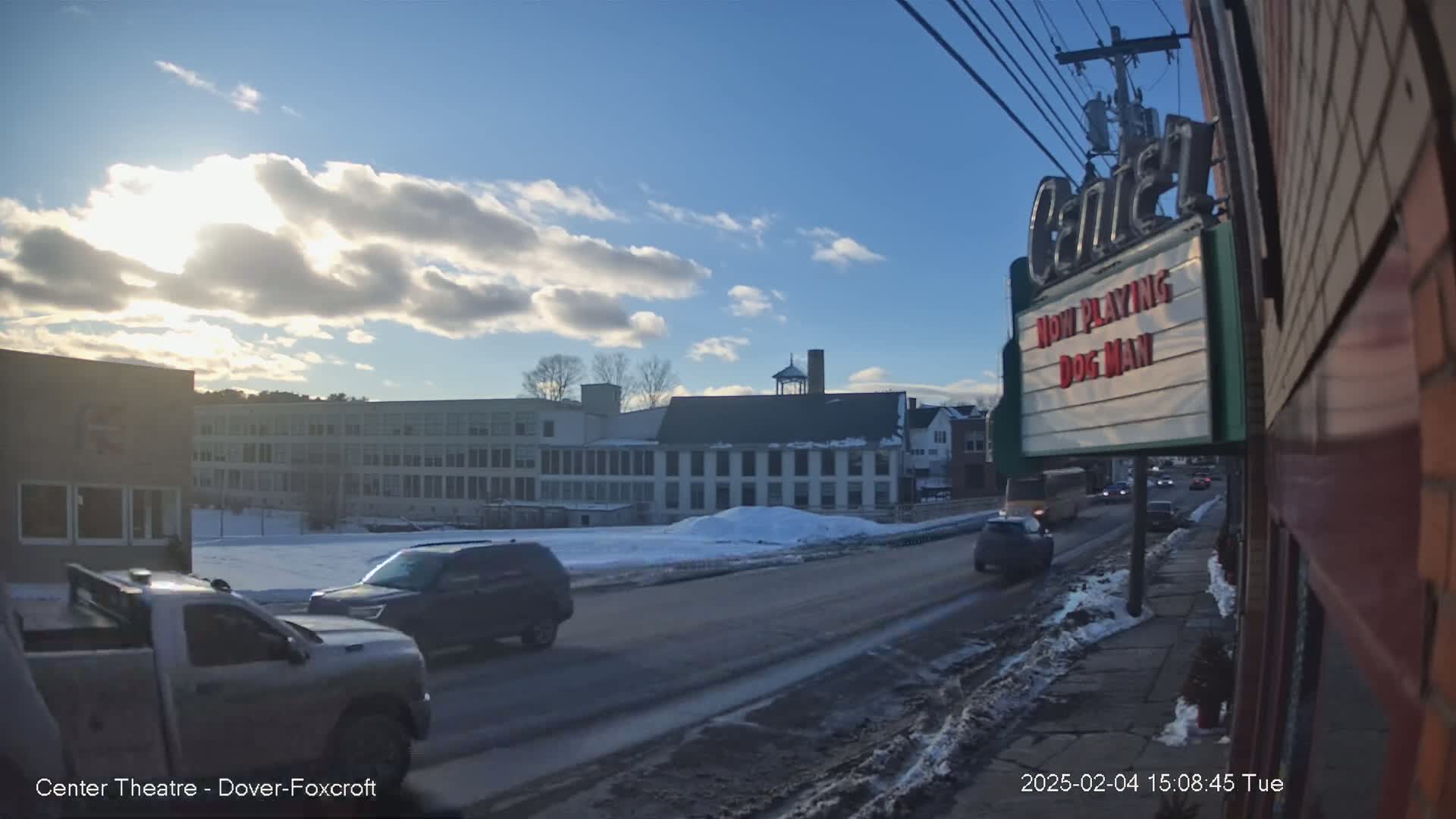 A sunny day with partly cloudy skies shows a street scene with several vehicles, including a pickup truck and SUV, driving past a multi-story building and a movie theater marquee.