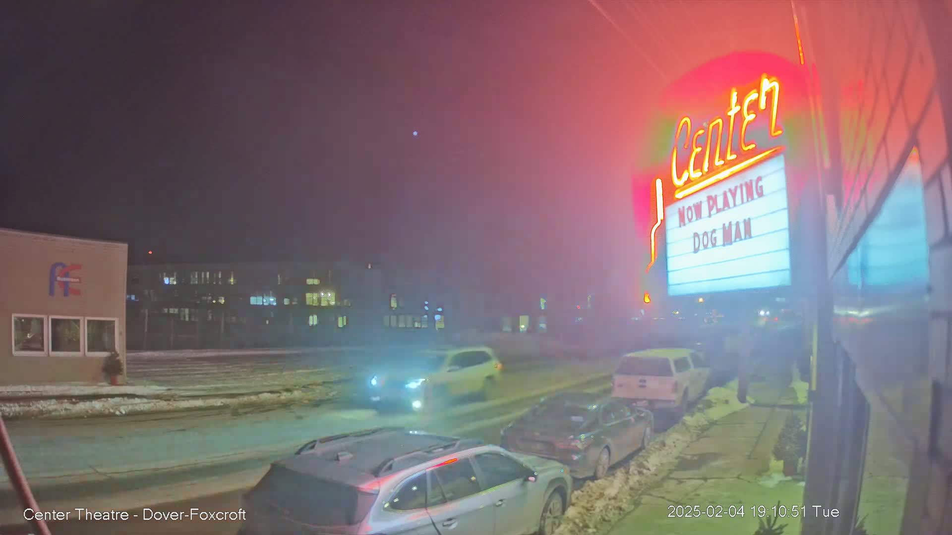 A hazy, snowy night scene shows several cars parked along a street in front of a movie theater marquee advertising "Dog Man."