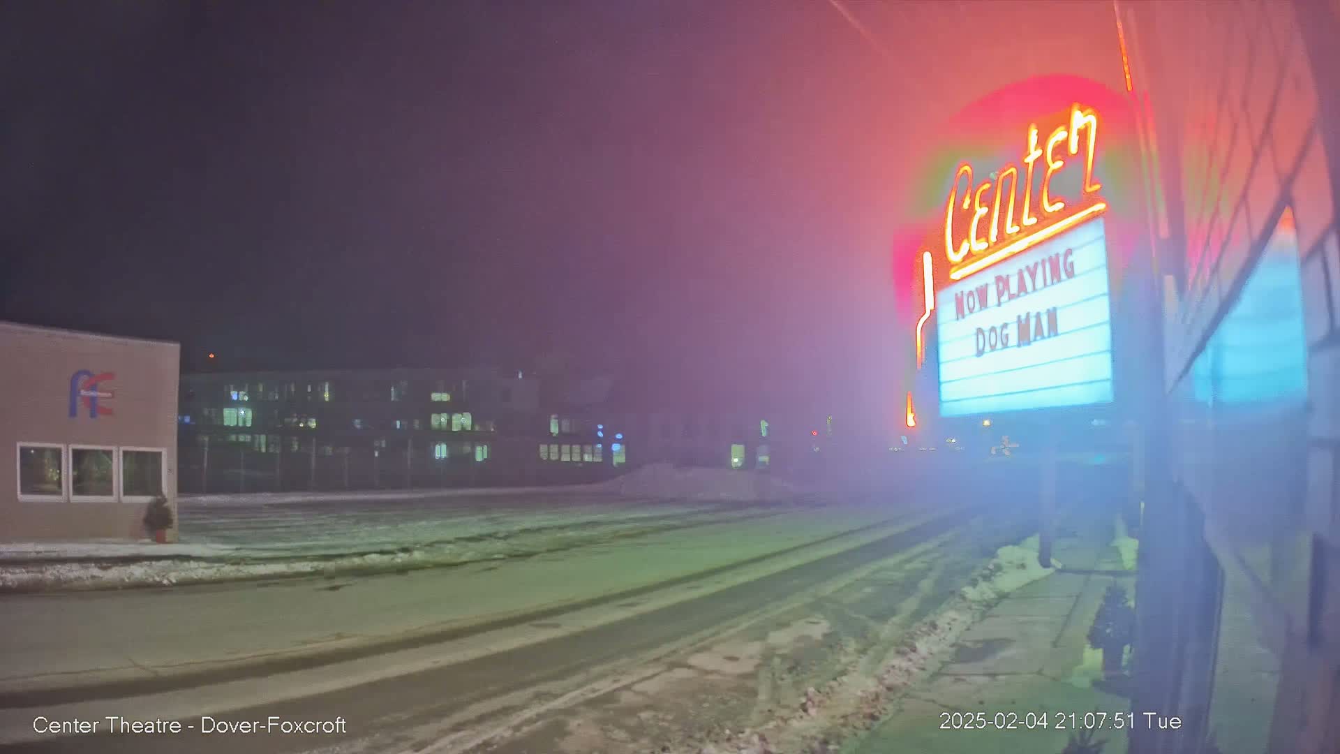 A snowy, foggy night scene shows a movie theater marquee advertising "Dog Man," with a building and a street visible in the background.
