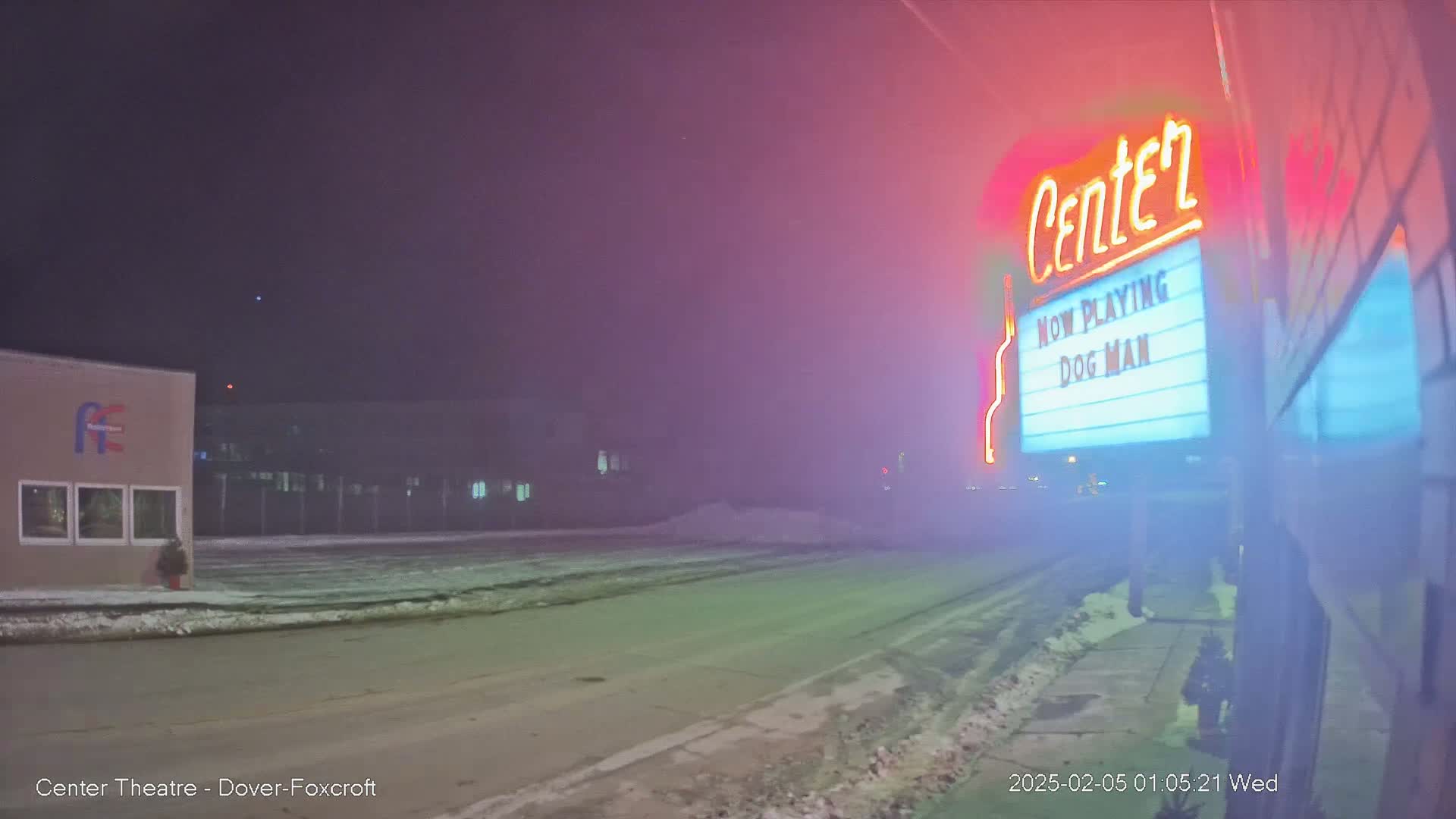 A foggy night scene shows a movie theater marquee advertising "Dog Man," with a snow-covered street and buildings in the background.