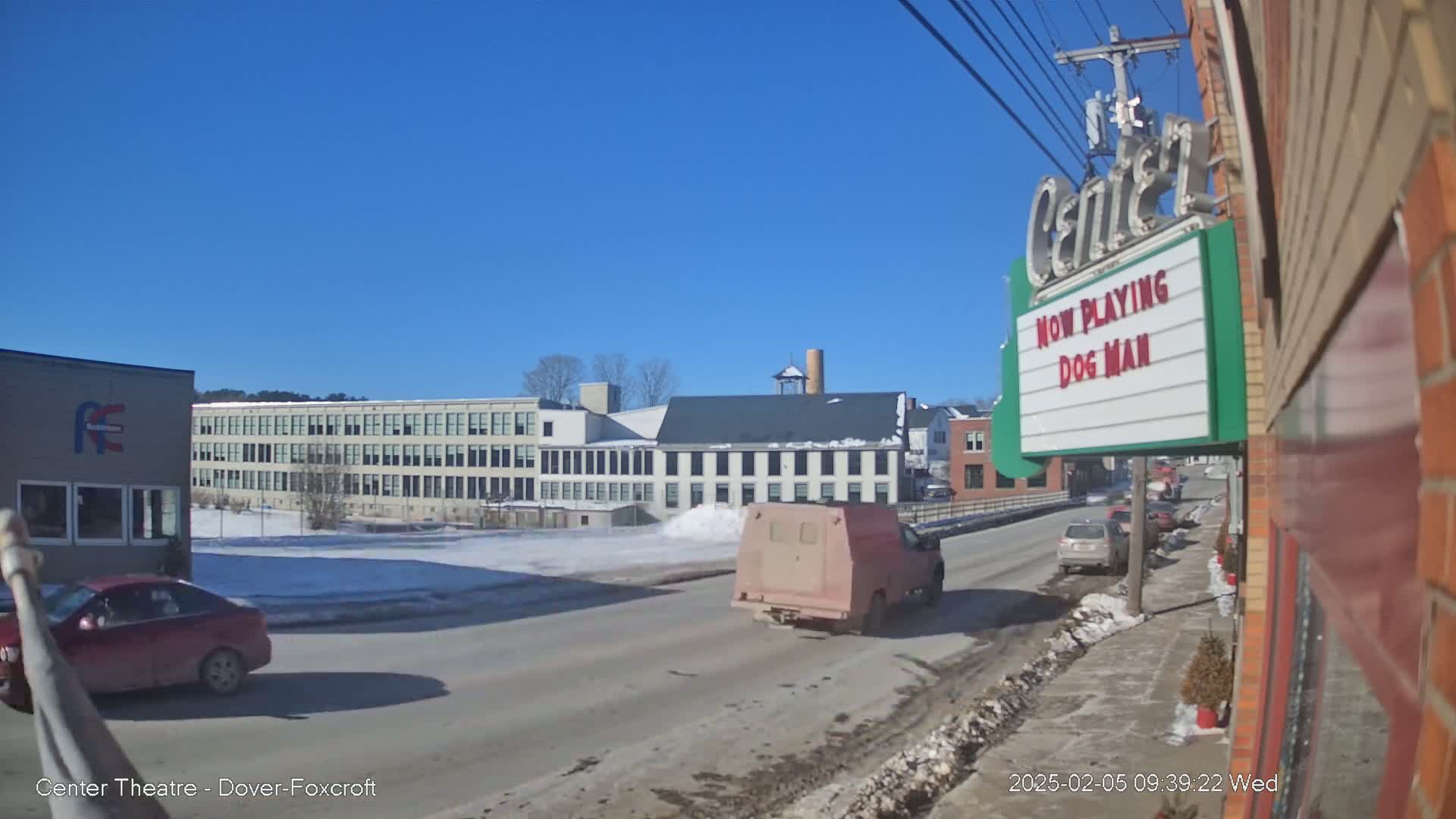A street scene on a sunny winter day shows a tan van and a red car driving past buildings, including a large factory and a movie theater marquee advertising "Dog Man."