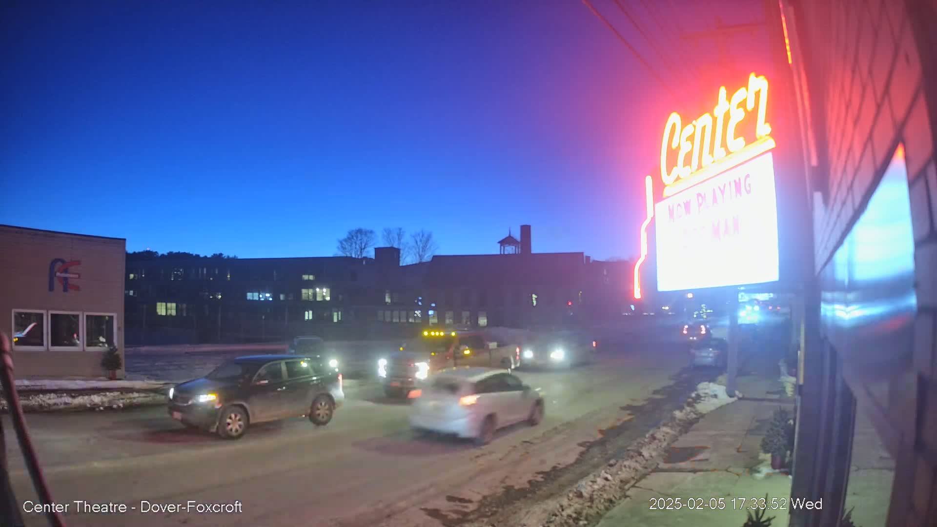 Several vehicles are driving on a snow-covered street at twilight, past a building with a neon sign that says "Center."