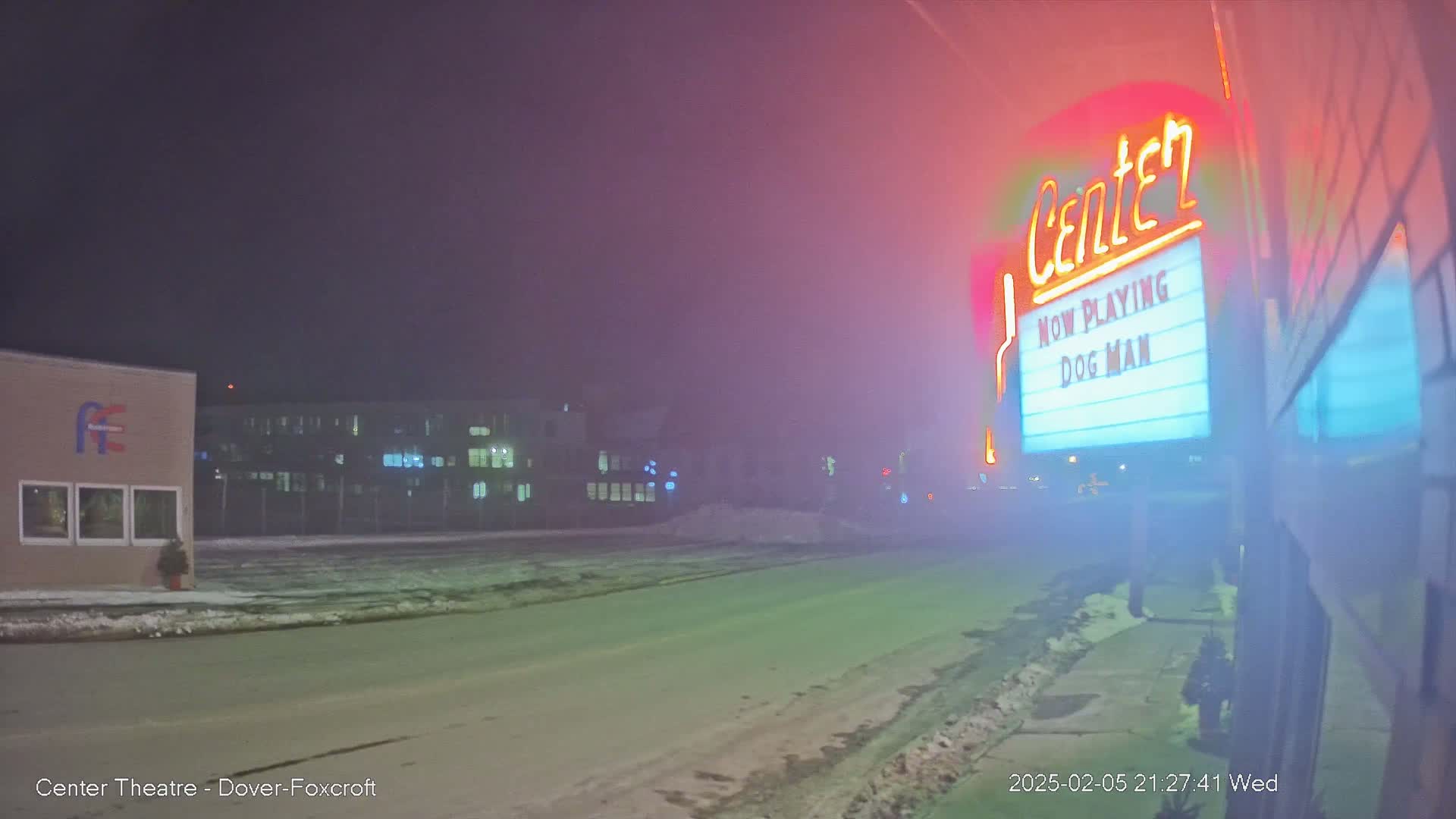 A foggy night scene shows a movie theater marquee advertising "Dog Man," with a snow-covered street and buildings in the background.