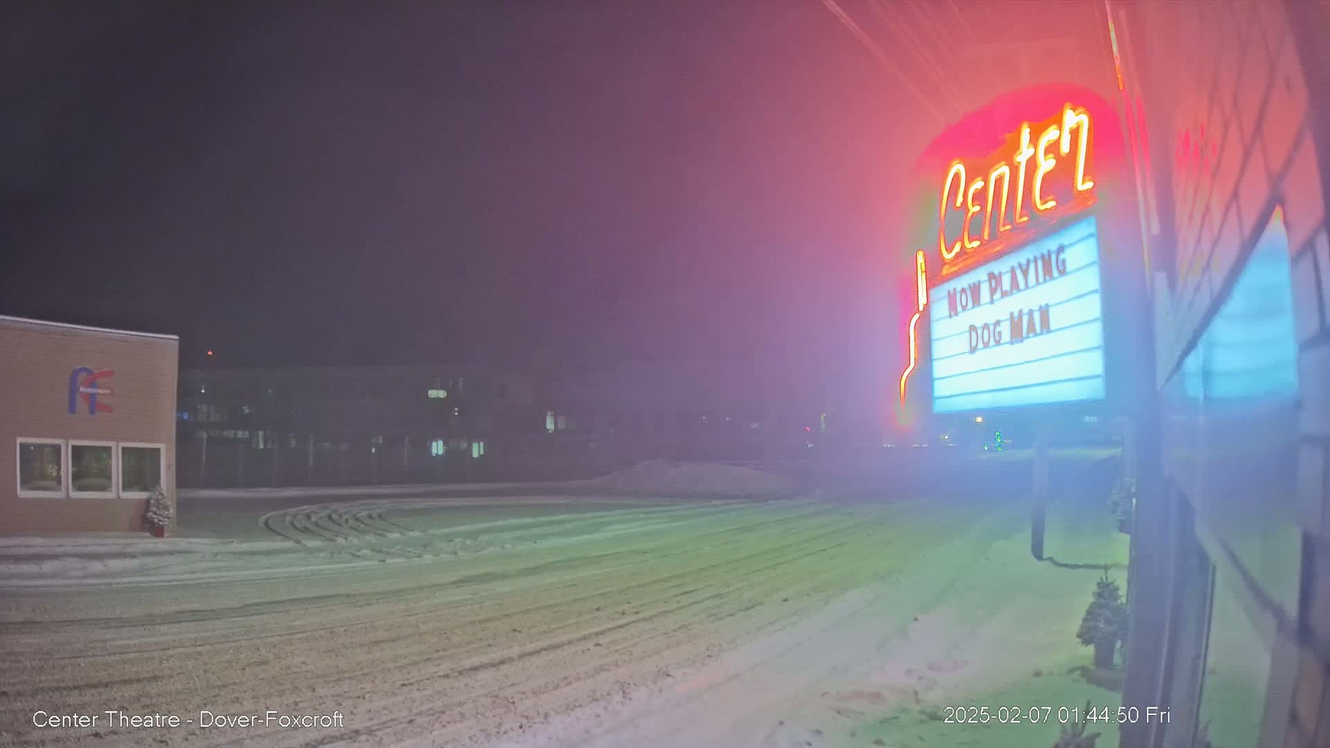 A snow-covered empty parking lot at night is seen in front of a movie theater marquee advertising "Dog Man".
