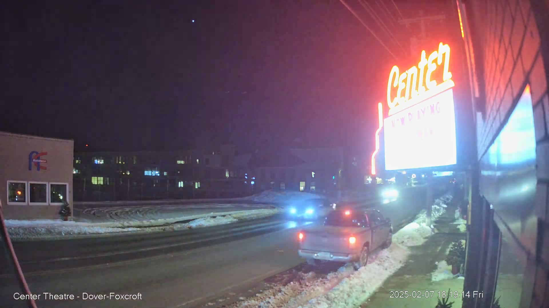 A snowy, nighttime street scene shows a pickup truck parked near a building with a lit-up "Center" sign.