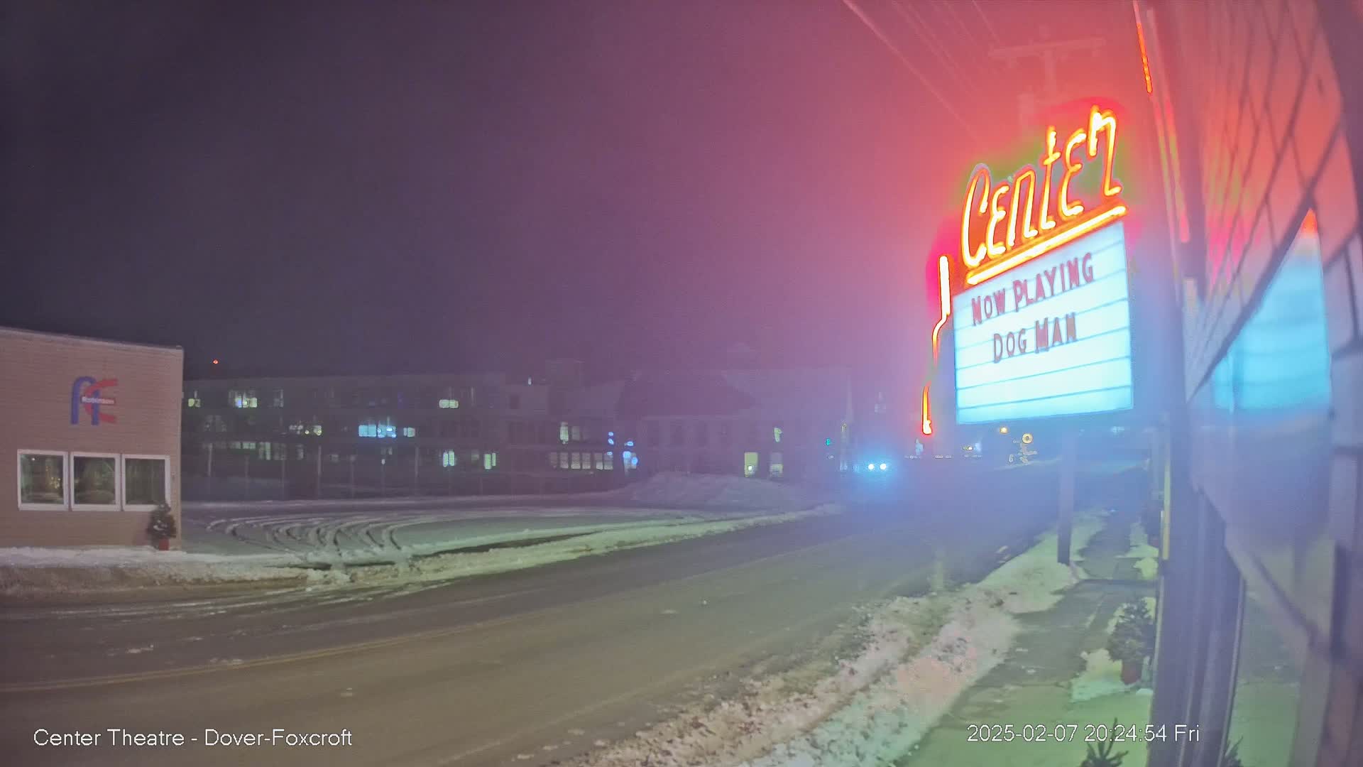 A snow-covered street at night is visible, with a movie theater marquee displaying "Dog Man" in the foreground, and a hazy atmosphere obscuring buildings in the background.