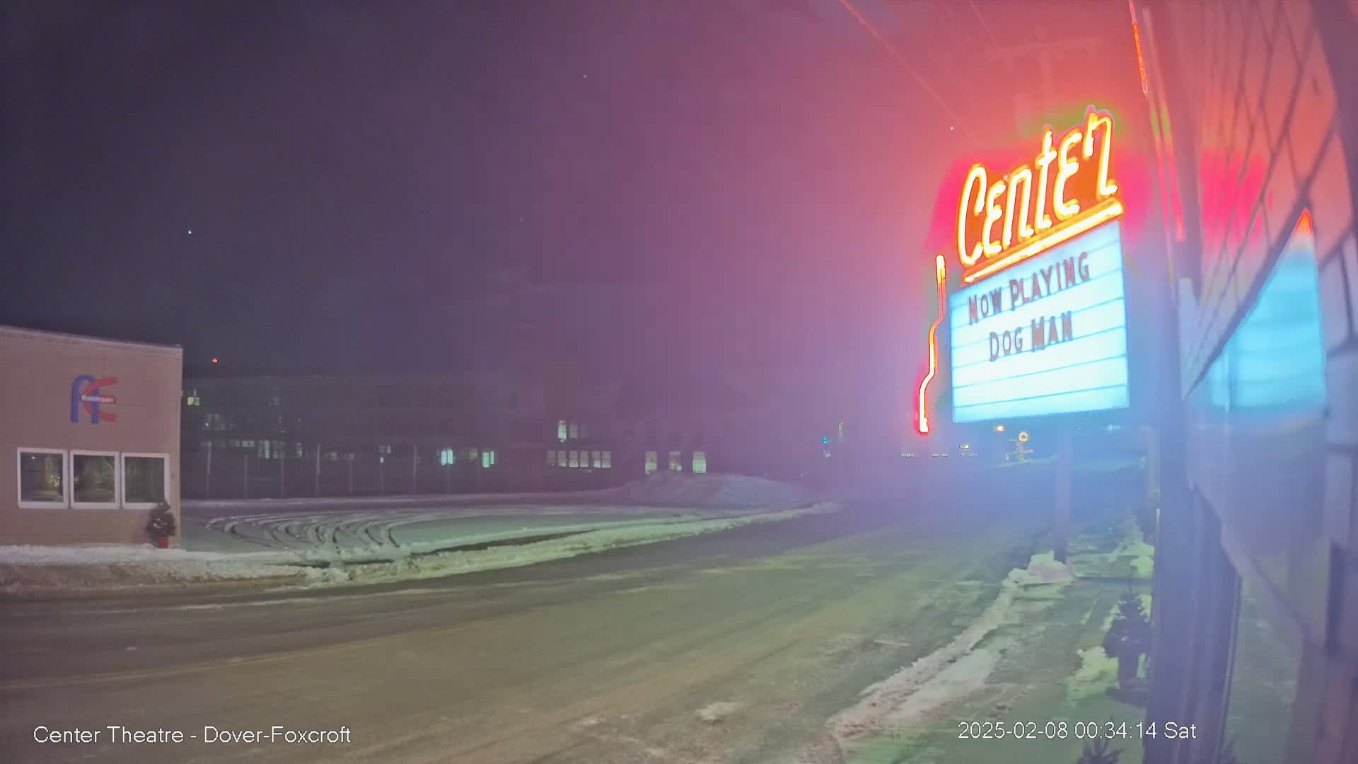 A snow-covered street at night, dimly lit by a neon movie theater marquee advertising "Dog Man."