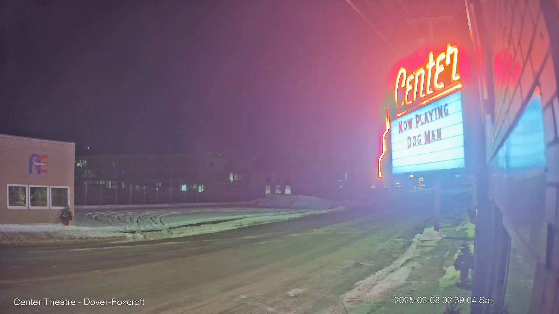 A snowy, hazy night scene shows a movie theater marquee advertising "Dog Man," with a building and street visible in the background.