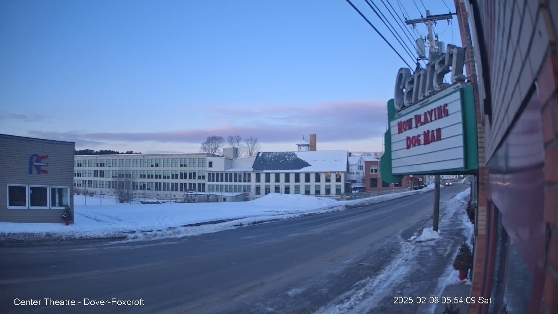 A snow-covered street scene at dawn shows several buildings, including a large multi-story structure and a movie theater marquee.