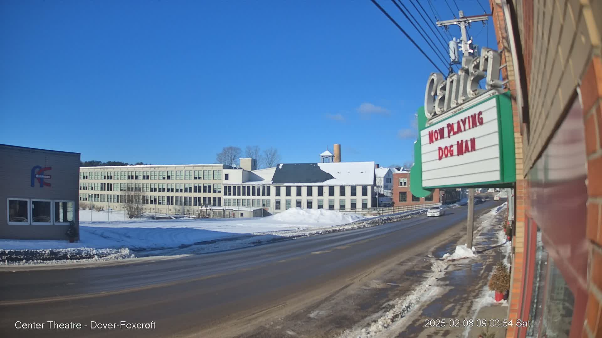 A snow-covered street scene on a sunny day shows several industrial buildings and a movie theater marquee.