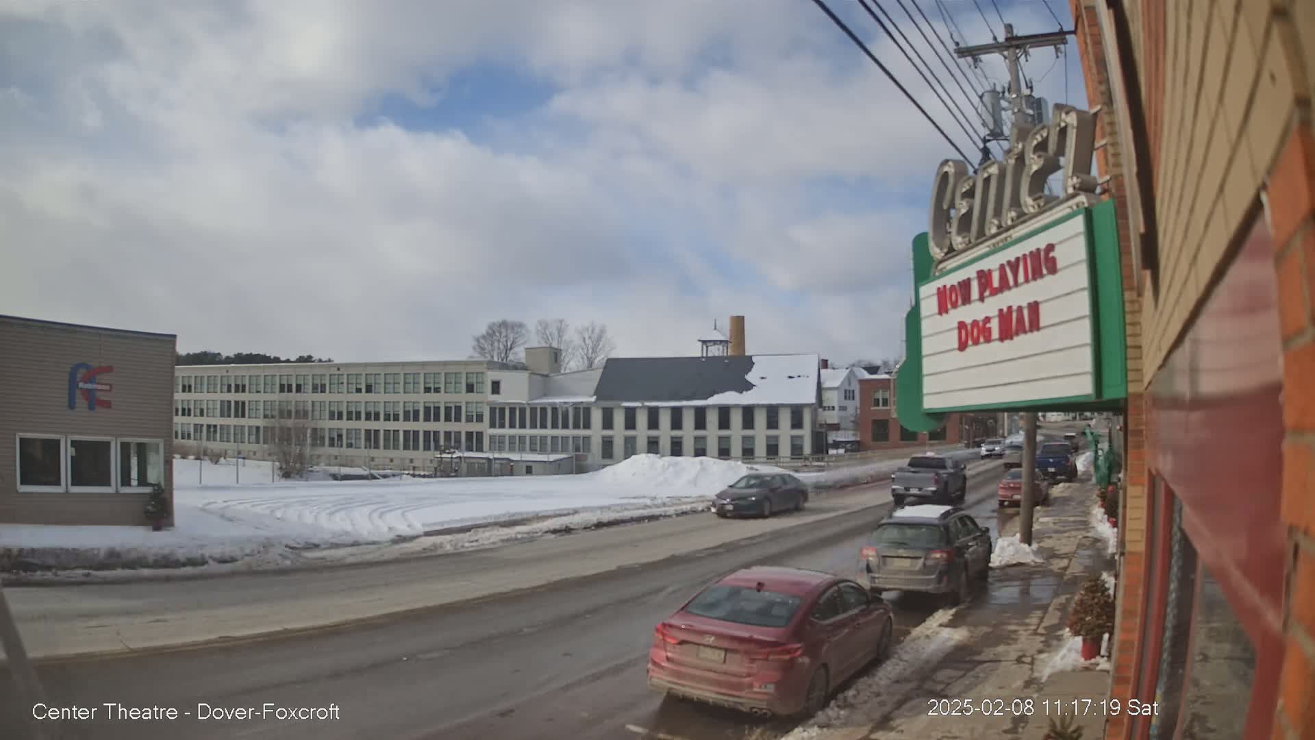 A partly cloudy winter day shows a street scene with several cars, a snow-covered landscape, and a large building in the background.