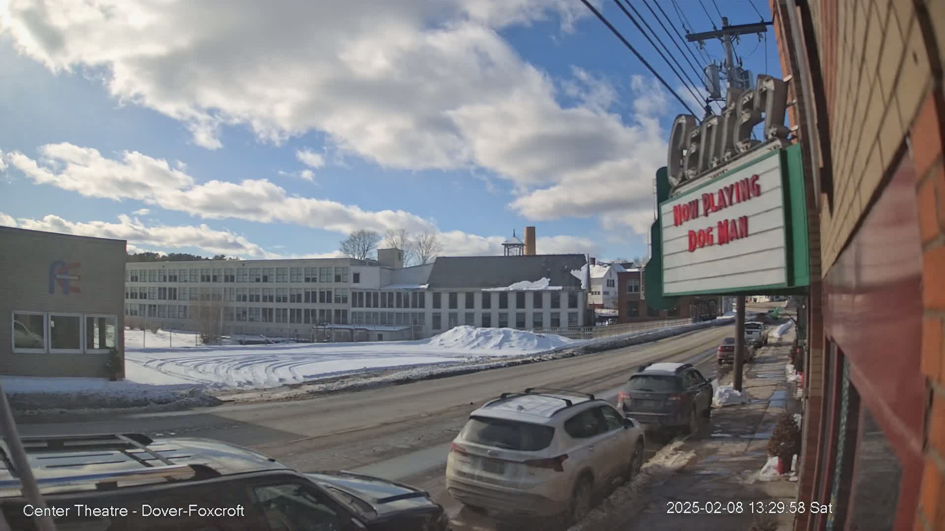 A partly sunny winter day reveals a street scene with cars parked along the side of a road, passing in front of buildings including a large multi-story structure and a movie theater marquee.