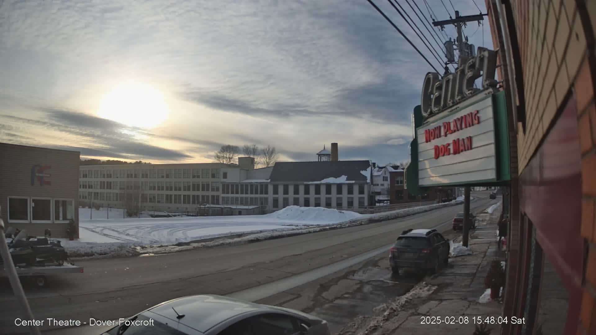 A partly sunny winter day shows a snow-covered street scene with several buildings, including a multi-story factory and a movie theater marquee.