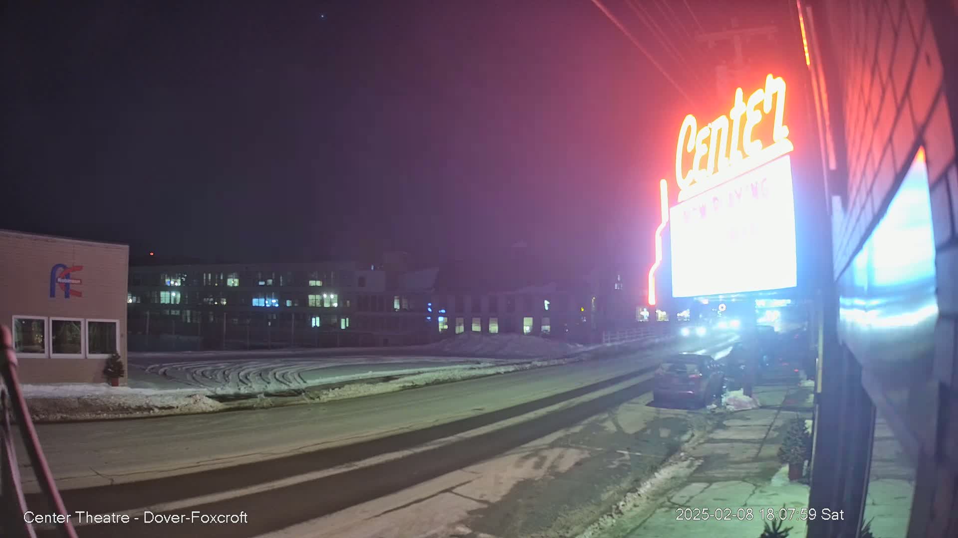 A snow-covered street at night leads to a building with a neon "Center" sign, and other buildings are visible in the background under a dark sky.