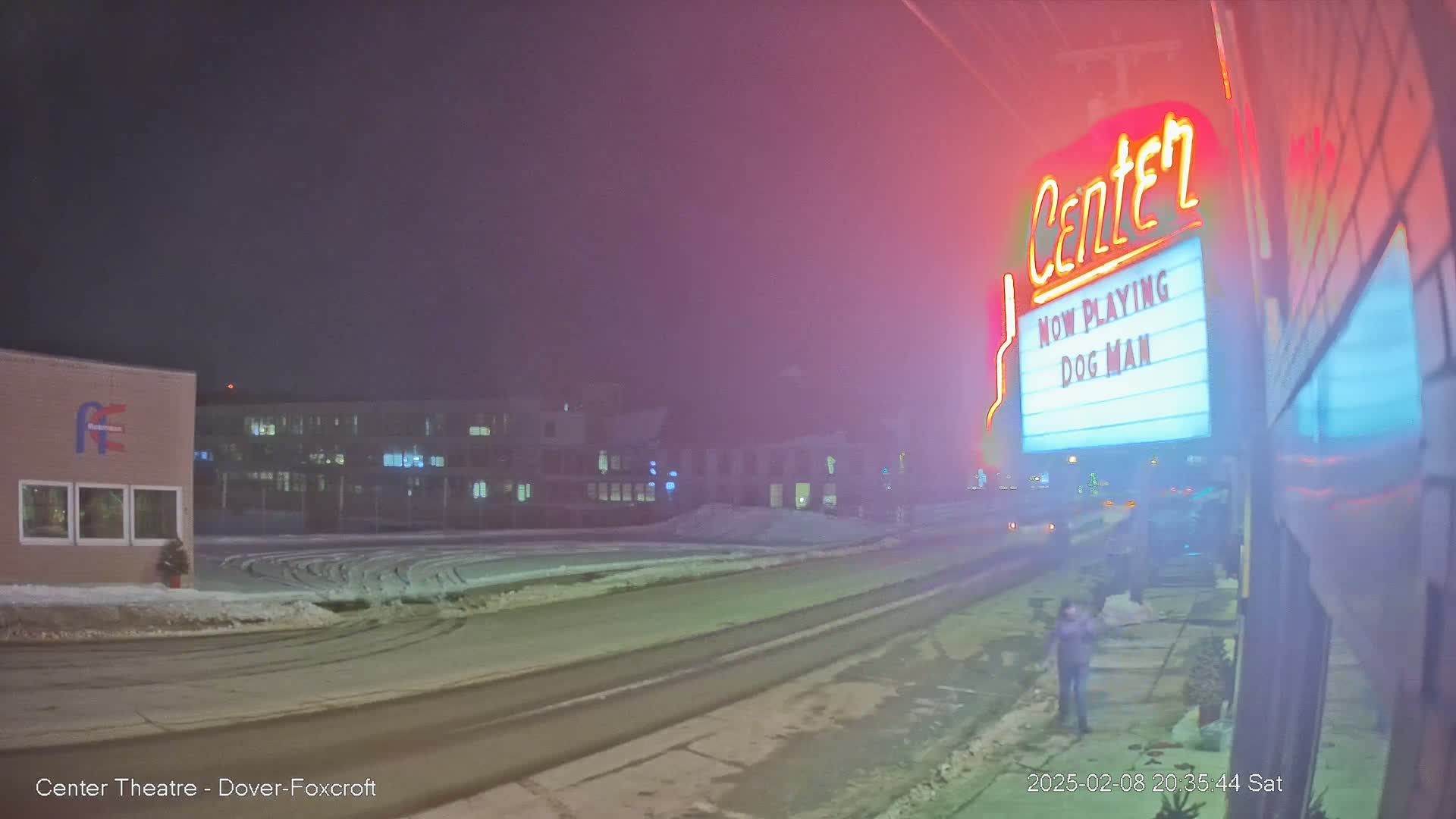 A snow-covered street at night shows a movie theater marquee advertising "Dog Man," with a hazy or foggy atmosphere.