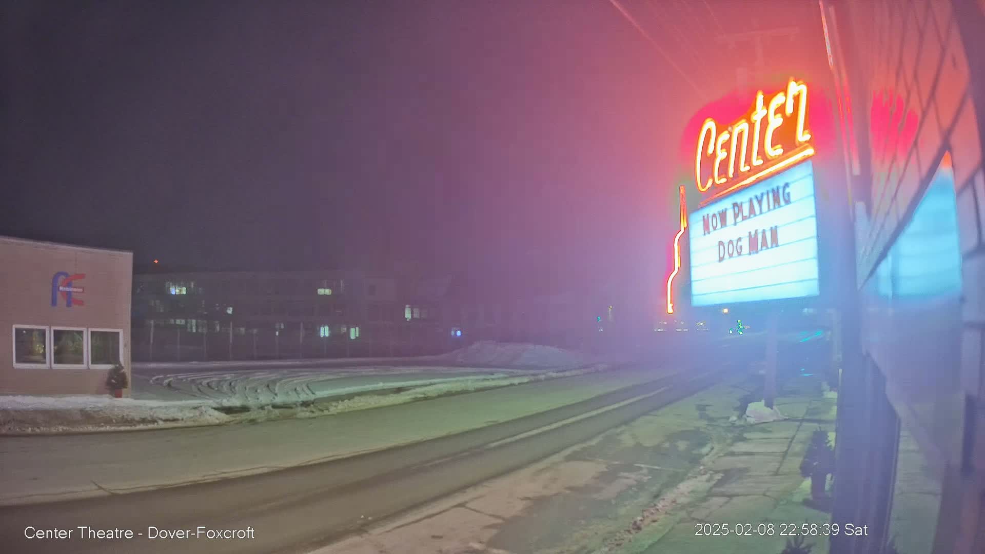 A foggy night scene shows a movie theater marquee advertising "Dog Man," with a snow-covered street and buildings in the background.