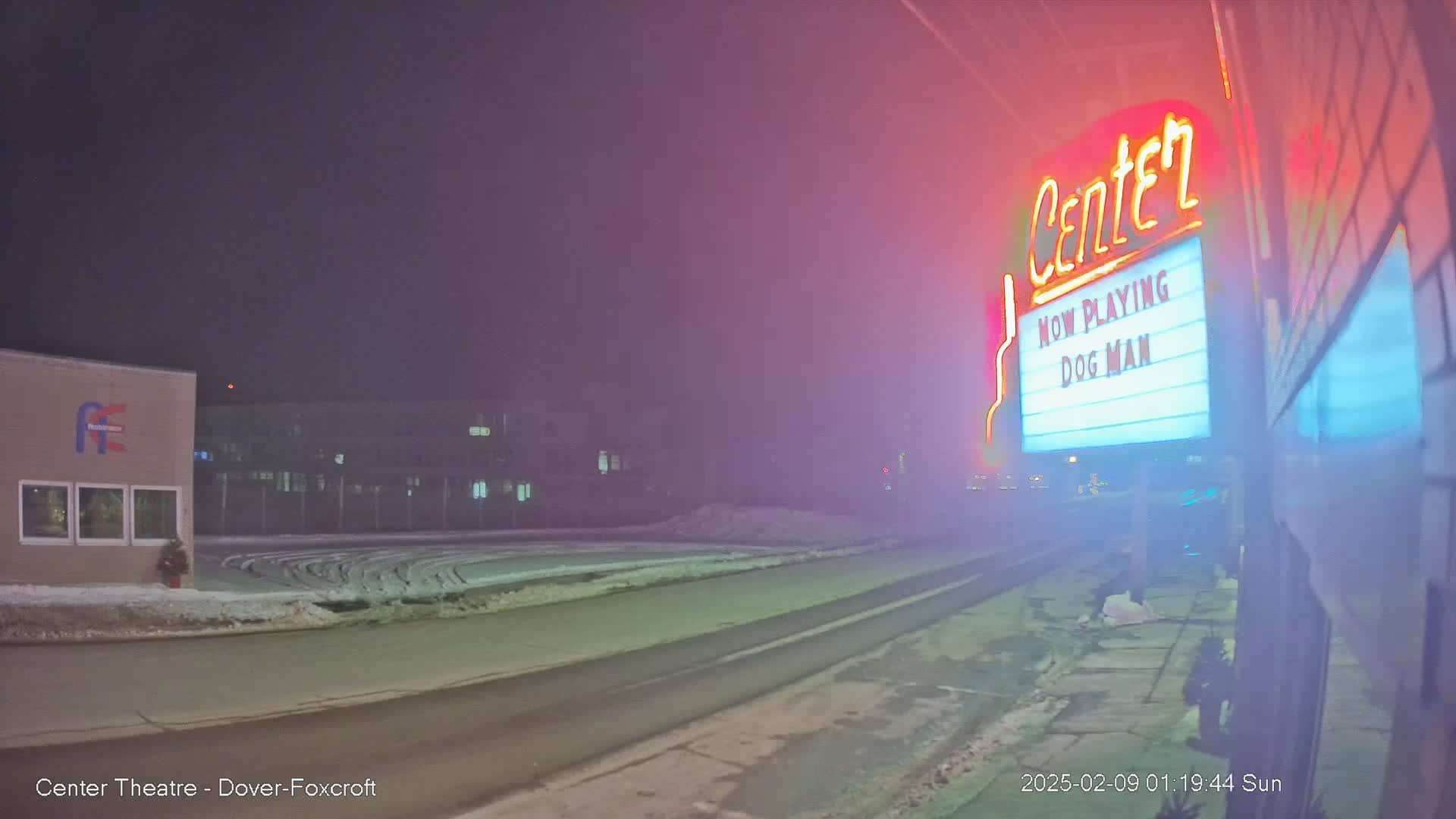A foggy night scene shows a road leading to a movie theater marquee displaying "Dog Man," with a snow-covered area and buildings in the background.