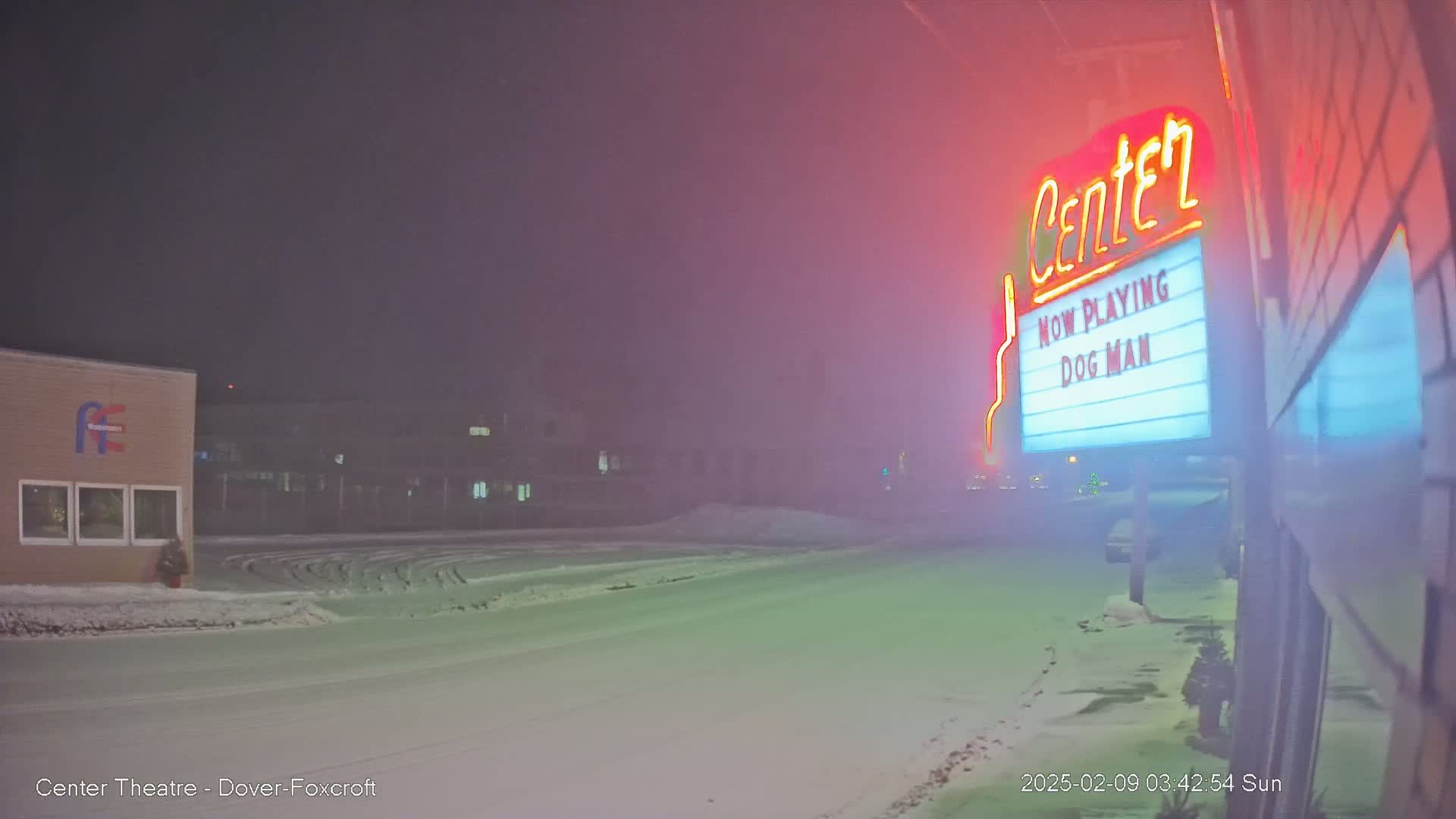 A snow-covered street at night, with a movie theater marquee advertising "Dog Man" visible in the fog.