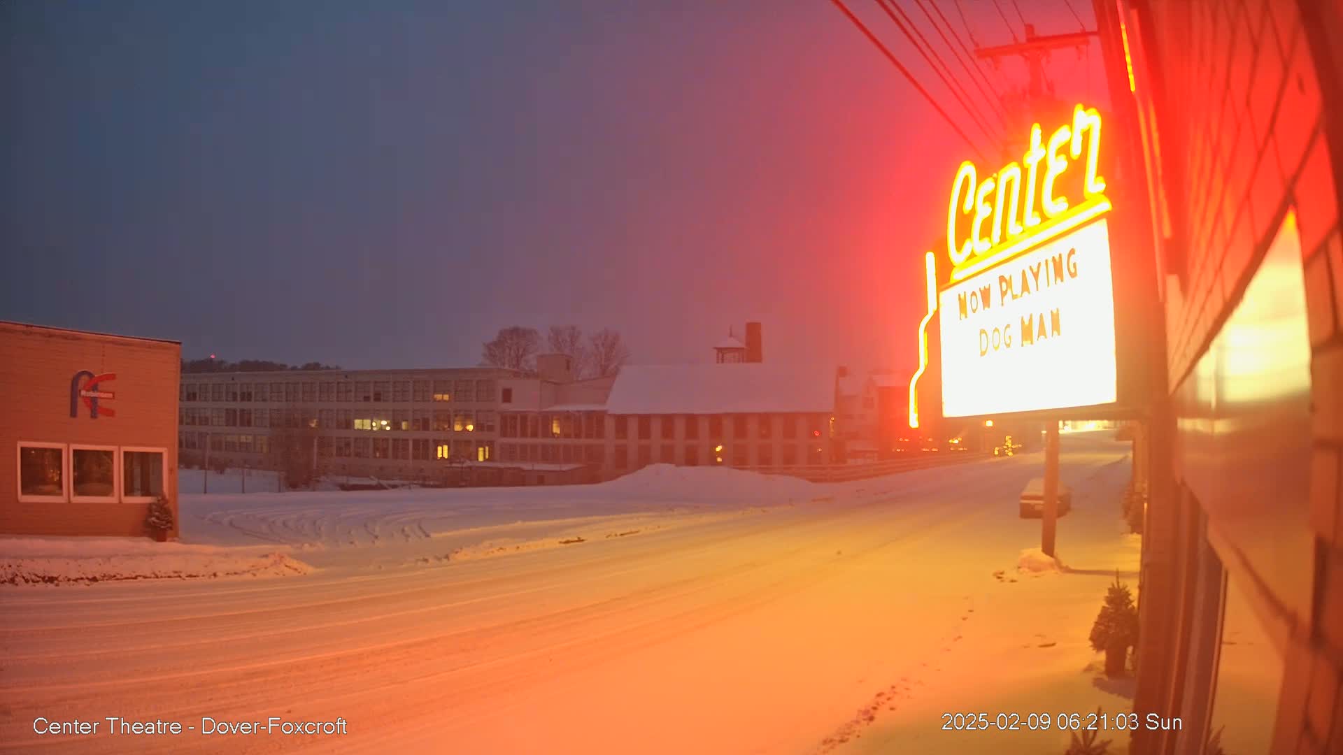 A snow-covered street at twilight leads toward buildings, one of which is a theater displaying a marquee.