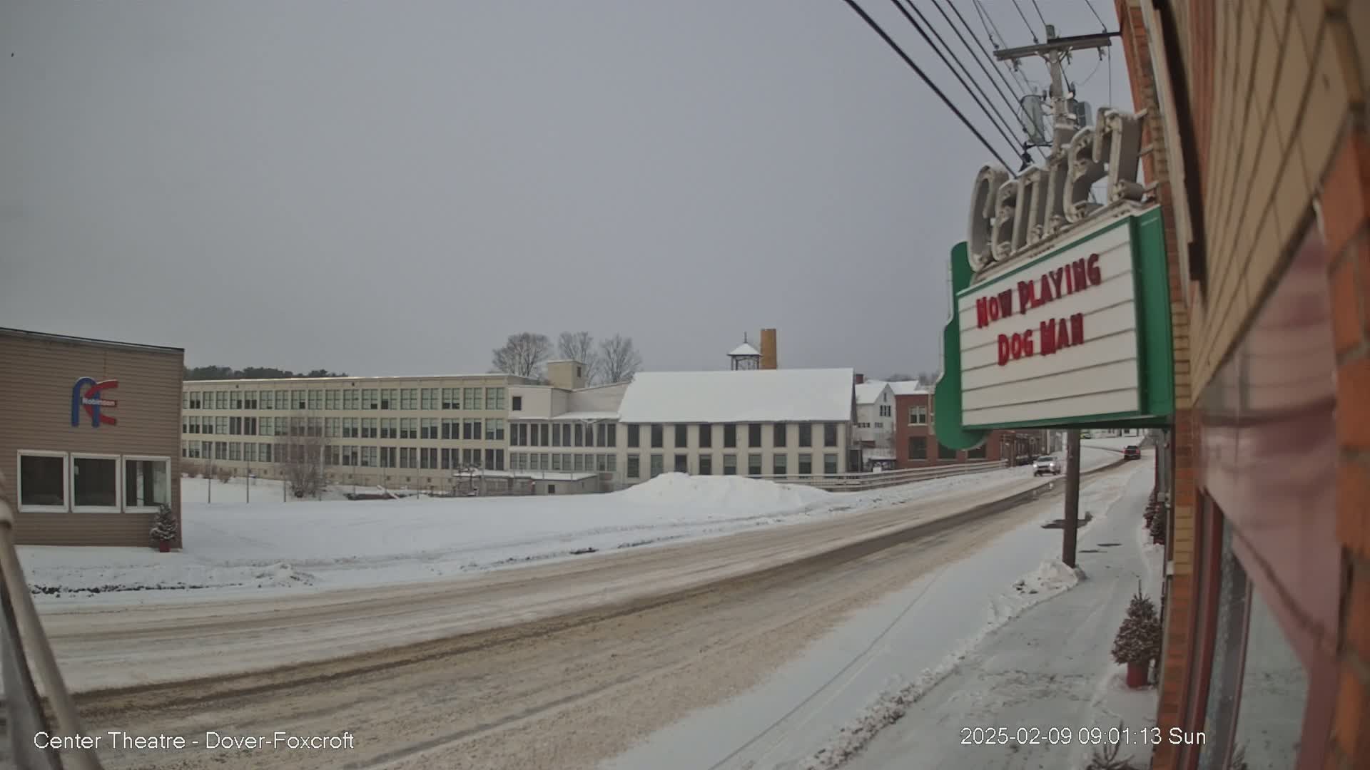A snow-covered street scene shows several buildings, including a large multi-story building and a smaller building with a marquee that says "NOW PLAYING DOG MAN".