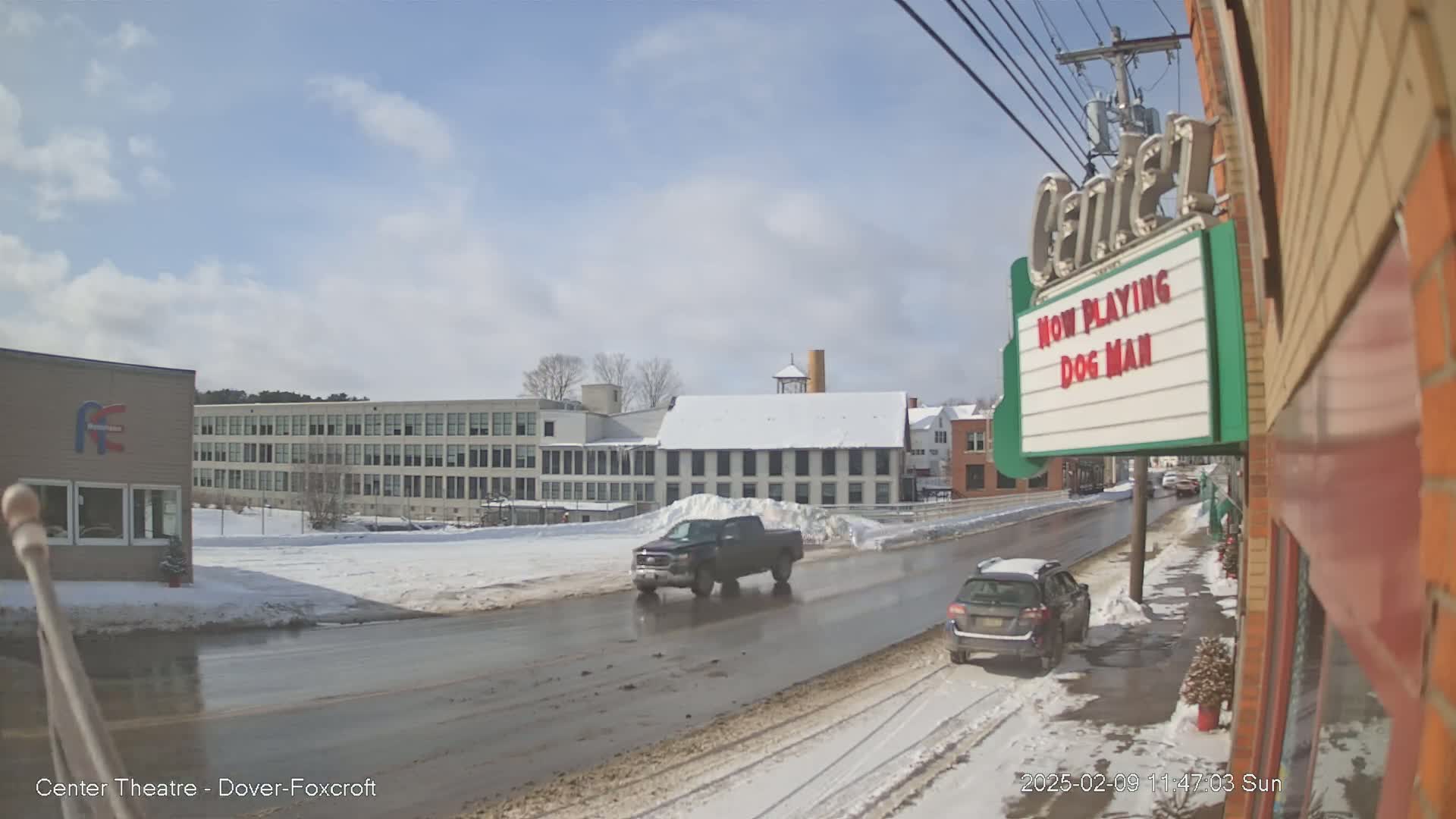 A partly cloudy winter day shows a street scene with a pickup truck and SUV driving on a wet road, past several buildings including a large factory and a movie theater showing "Dog Man."