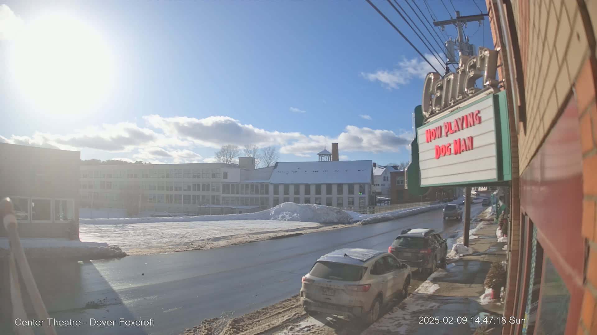 A sunny winter day shows a snow-covered street with several cars parked along the side, and buildings in the background.