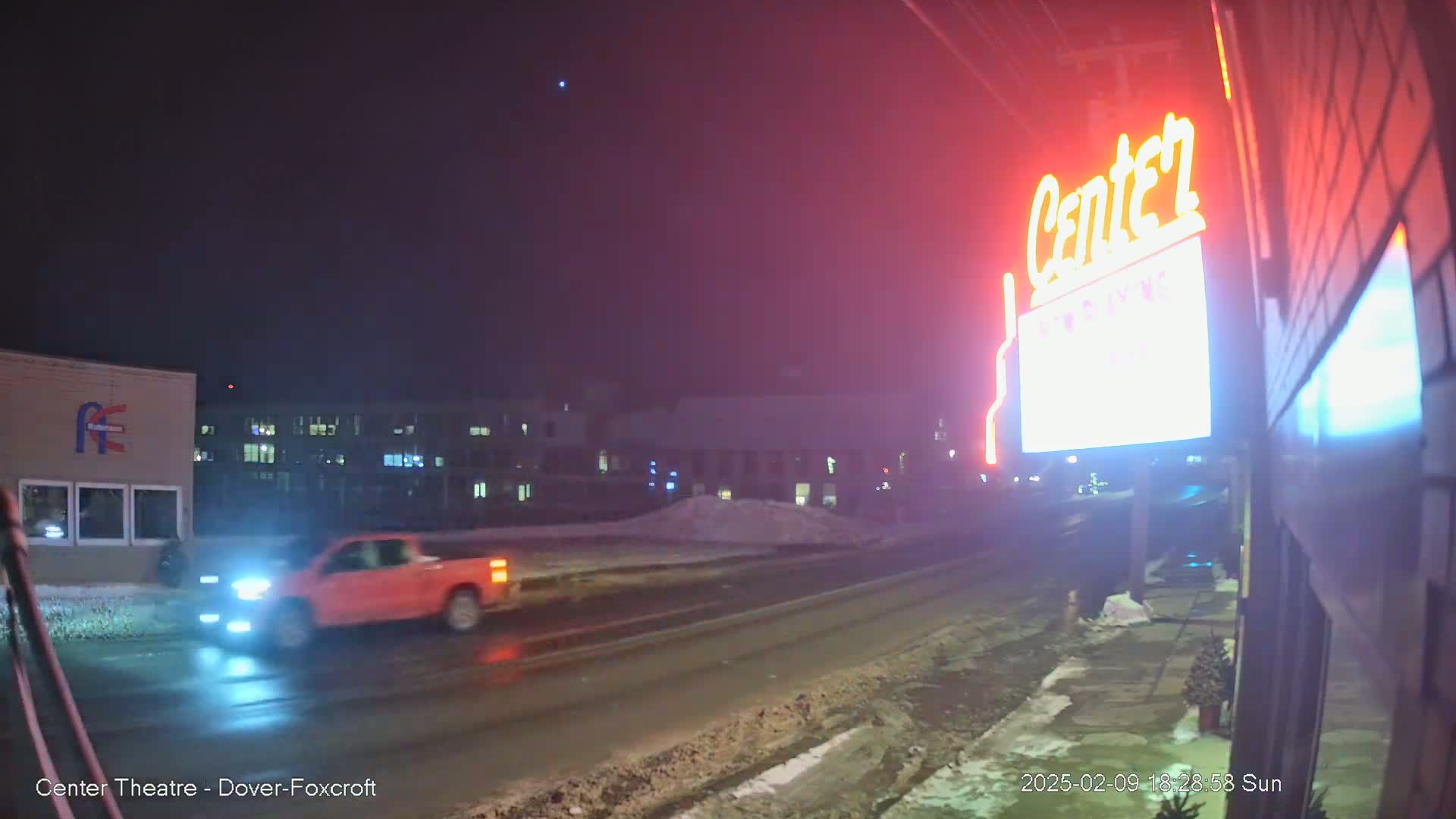 A red pickup truck drives past a brightly lit "Center" sign on a snowy, nighttime street.